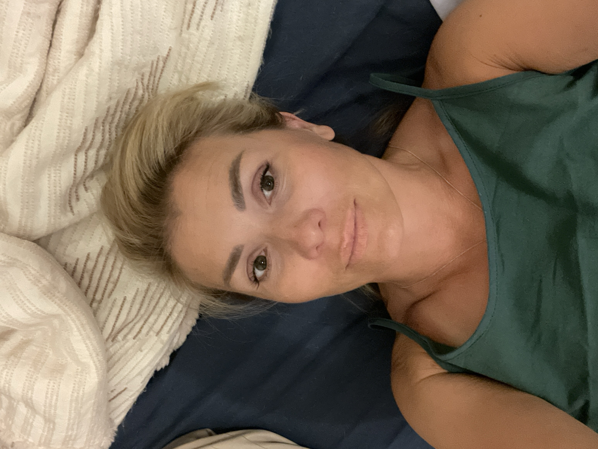 A young, Caucasian woman with blonde hair is lying down, looking at the camera. She is wearing a dark green spaghetti strap top. Her head is tilted to the right, and her eyes are open and looking directly at the viewer. Her hair is pulled back, revealing her face. She appears to be in a relaxed and casual setting, possibly in bed, as she is lying on a dark blue surface that resembles a mattress, with a cream-colored blanket on the left side of the image. The lighting suggests it could be daytime, but the exact time of day is not discernible. There is no visible text in the image. The woman's expression is neutral, with a slight, subtle smile.