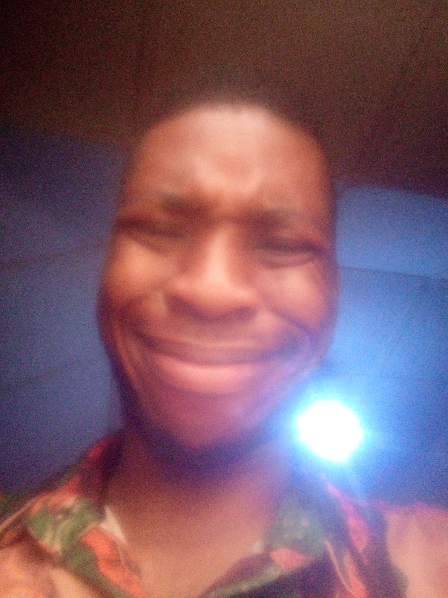 A close-up, blurry photograph shows a person with dark skin and short hair. The person's eyes are partially closed in a smile. A bright, circular light source obscures part of the person's face. The person is wearing a collared shirt with a colorful pattern. The background is dark and indistinct. The location is unknown.