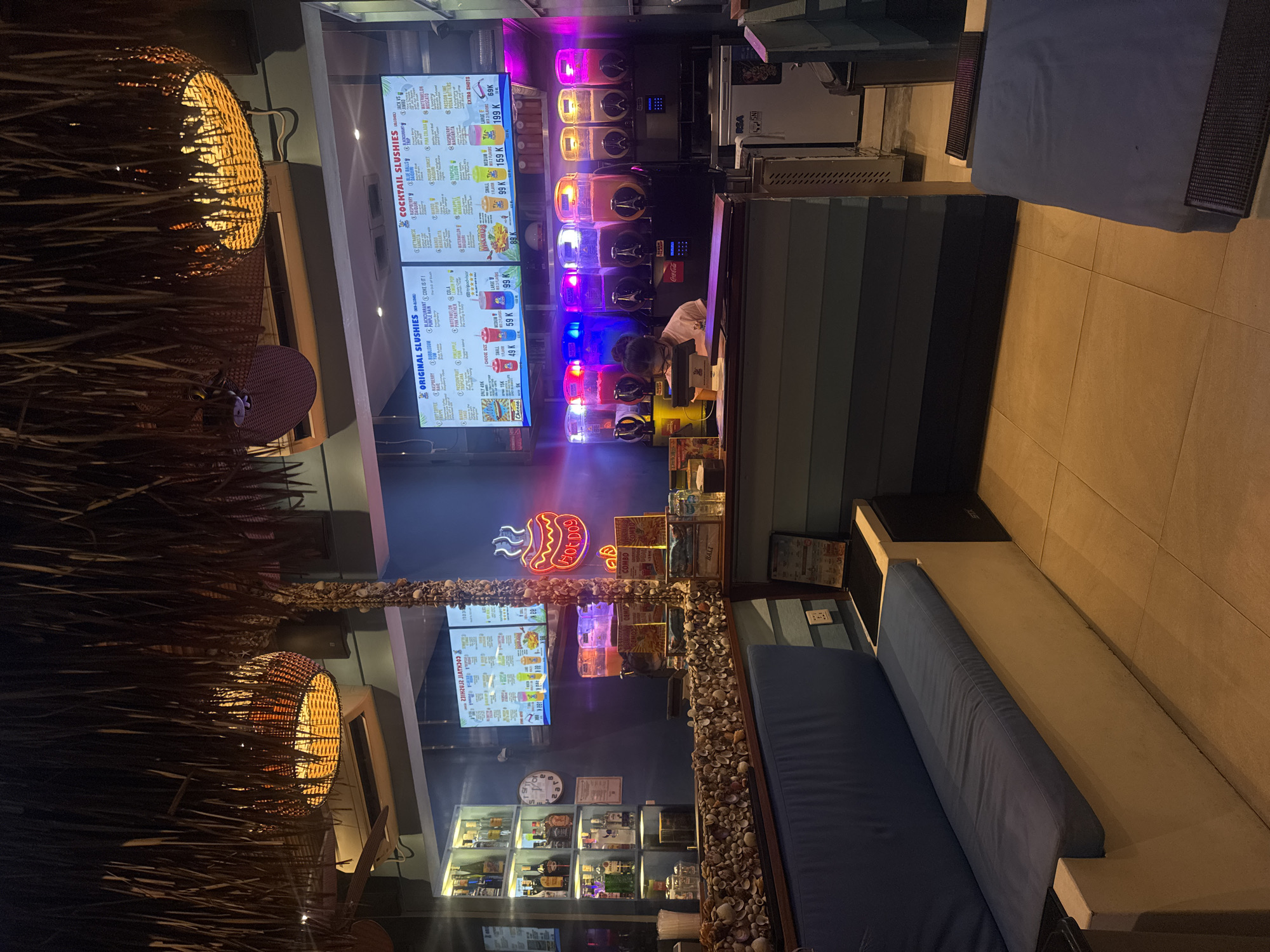 The image depicts the interior of a bar or cafe in Kuta, Indonesia, illuminated by ambient lighting and neon signs. A thatched roof overhang with a woven basket light fixture is visible in the foreground. Behind a counter adorned with seashells, several slushie machines are dispensing colorful beverages. A person wearing a pink shirt and glasses is seated at the counter, appearing to be working. A digital menu board displaying "ORIGINAL SLUSHIES" and "COCKTAIL SLUSHIES" with prices is mounted above the machines. A neon "HOT DOG" sign glows in the background. The environment suggests a casual establishment, possibly a beachside bar, with seating areas furnished with blue cushions.