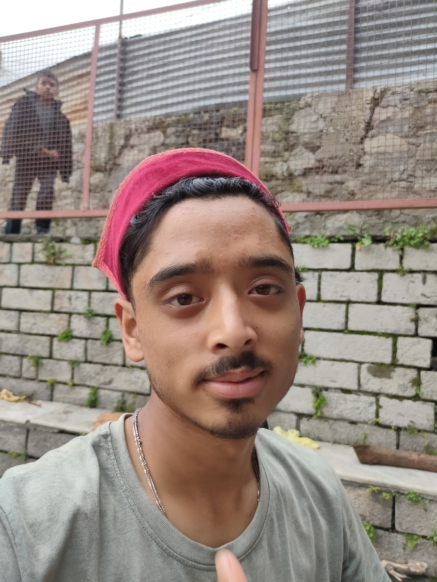 A young man wearing a red bandana and a light green t-shirt is posing for a selfie. He has short, dark hair and a small mustache and goatee. A thin silver chain is visible around his neck. In the background, a person wearing a dark jacket and pants is standing behind a metal fence. The background also features a stone wall and some greenery. The scene appears to be outdoors in an urban or semi-urban environment.