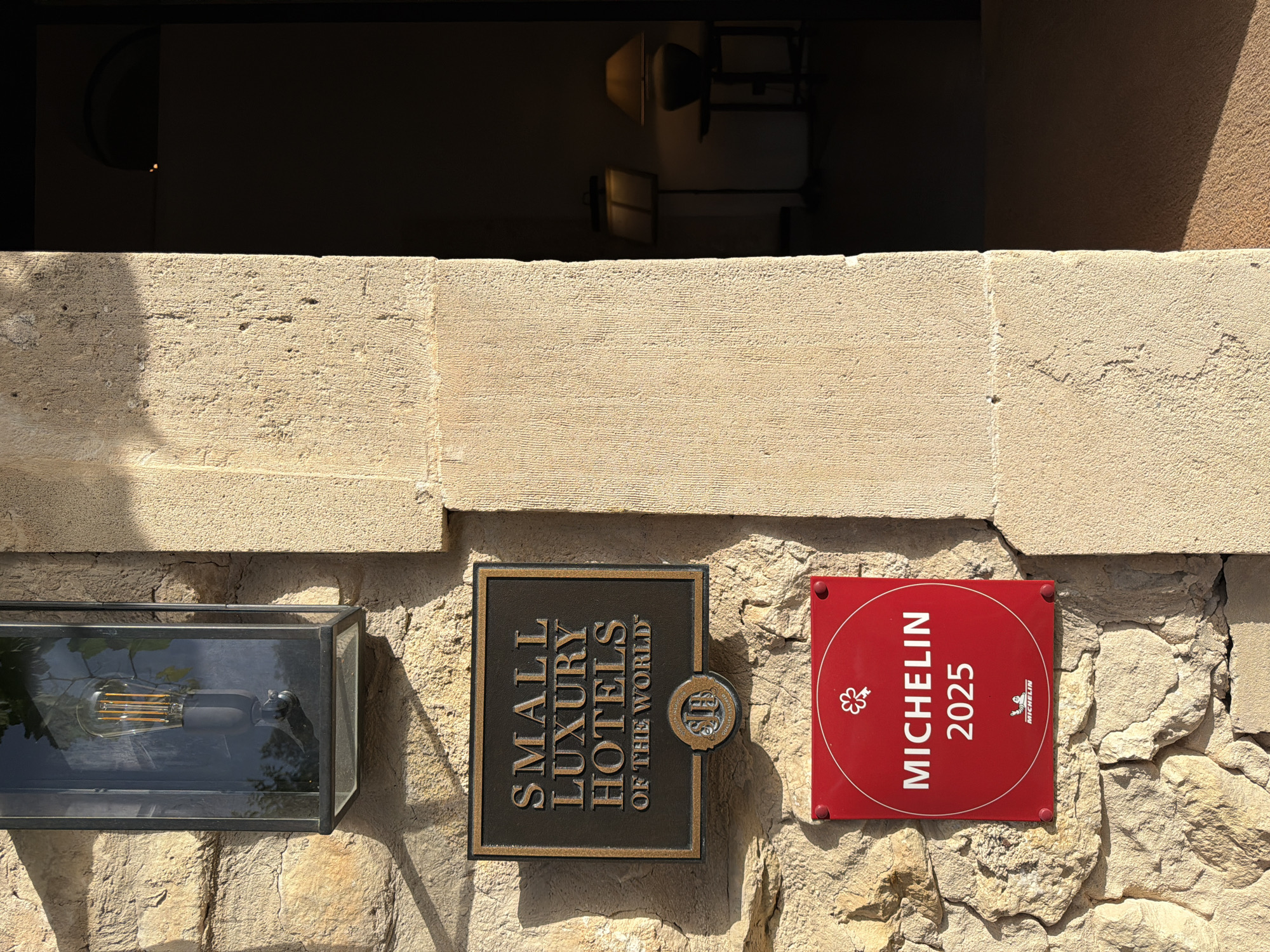 This image depicts a close-up view of a stone wall on the exterior of a building, likely a hotel, in Montuïri, Spain. Two prominent signs are mounted on the wall. The upper sign is a dark, rectangular plaque with gold lettering that reads "SMALL LUXURY HOTELS OF THE WORLD" and a circular emblem below it. The lower sign is a square, vibrant red plaque with white lettering that says "MICHELIN 2025" and features a stylized key symbol at the top and a Michelin Man logo at the bottom. To the right of the signs, a shadowed doorway leads into a dimly lit interior where two lamps are visible. The stone wall itself is rough-hewn and bathed in warm sunlight, casting distinct shadows. An outdoor lantern with an exposed Edison bulb is mounted above the "SMALL LUXURY HOTELS" sign. The overall impression is one of a distinguished establishment with notable accreditations.