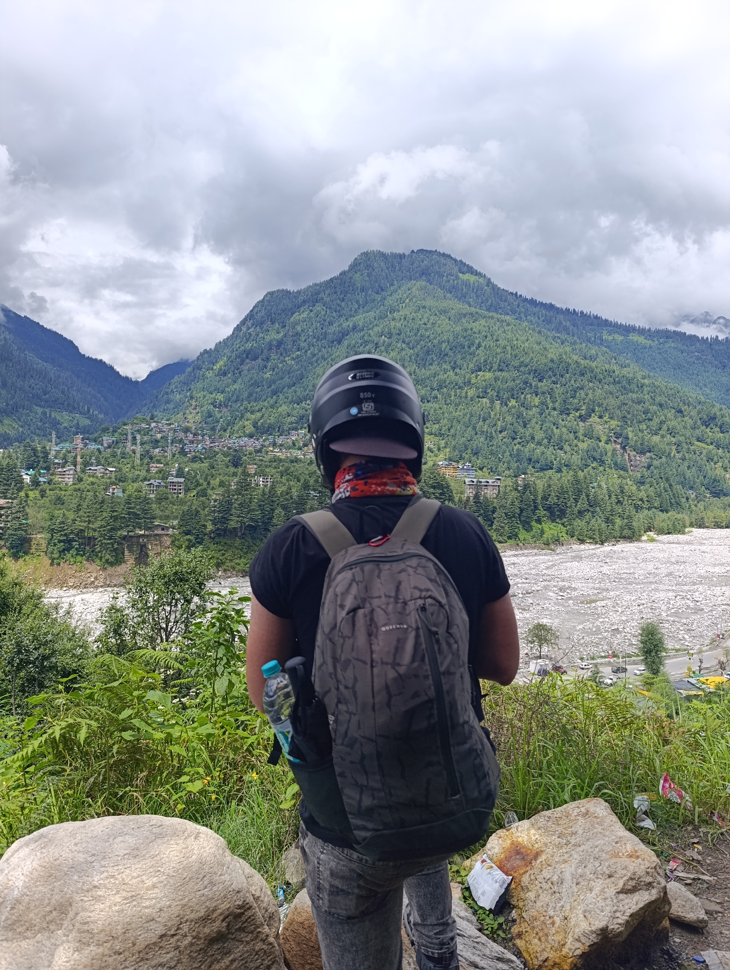 A person wearing a black t-shirt, gray jeans, and a black helmet stands with their back to the camera, facing a mountainous landscape in Manali, India. They are carrying a gray backpack with a water bottle attached to the side. In the background, a town with buildings is nestled on the side of a lush green mountain. A wide, rocky riverbed runs through the valley below, with a road and vehicles visible in the distance. The sky is overcast with clouds. The foreground features large rocks and dense green vegetation.