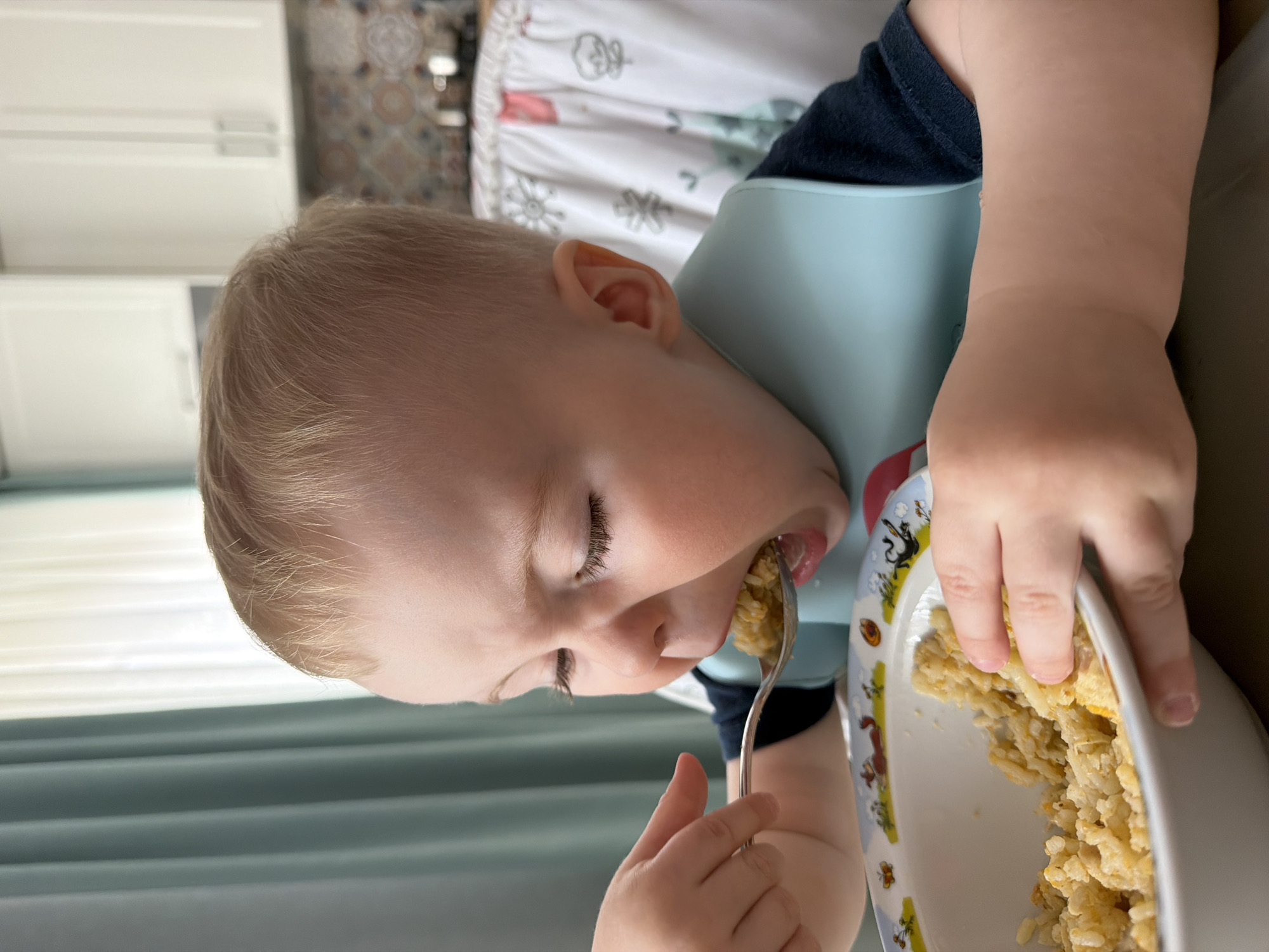 The image, rotated 90 degrees counter-clockwise, depicts a young child actively eating a meal. The child, with fair skin and light blonde hair, is shown in profile, with eyes closed in concentration, bringing a spoonful of food to their mouth. They are wearing a light blue silicone bib over a dark blue t-shirt. One hand holds a metal spoon filled with a yellowish-white, grain-based dish, likely rice, while the other hand rests on the edge of a white bowl. The bowl is adorned with colorful cartoon-like illustrations, including a black cat and a horse.
The setting appears to be a brightly lit indoor space, possibly a kitchen or dining area, suggesting it is daytime. In the background to the left, white kitchen cabinets are visible, along with a window covered by light teal horizontal blinds or curtains. To the right and behind the child, a patterned fabric, possibly part of a high chair or seating, can be seen, behind which is a tiled wall with decorative patterns. The child's focused expression suggests deep engagement with their meal. No discernible text is visible in the image.
