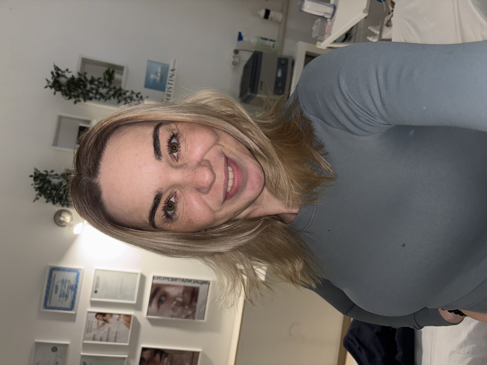 A selfie of a fair-skinned woman with blonde hair. She is wearing a gray long-sleeved shirt and smiling. Behind her is a wall with framed certificates and photographs. Greenery is visible on a shelf in the background. Medical equipment is also visible in the background. The city and location are unknown.
