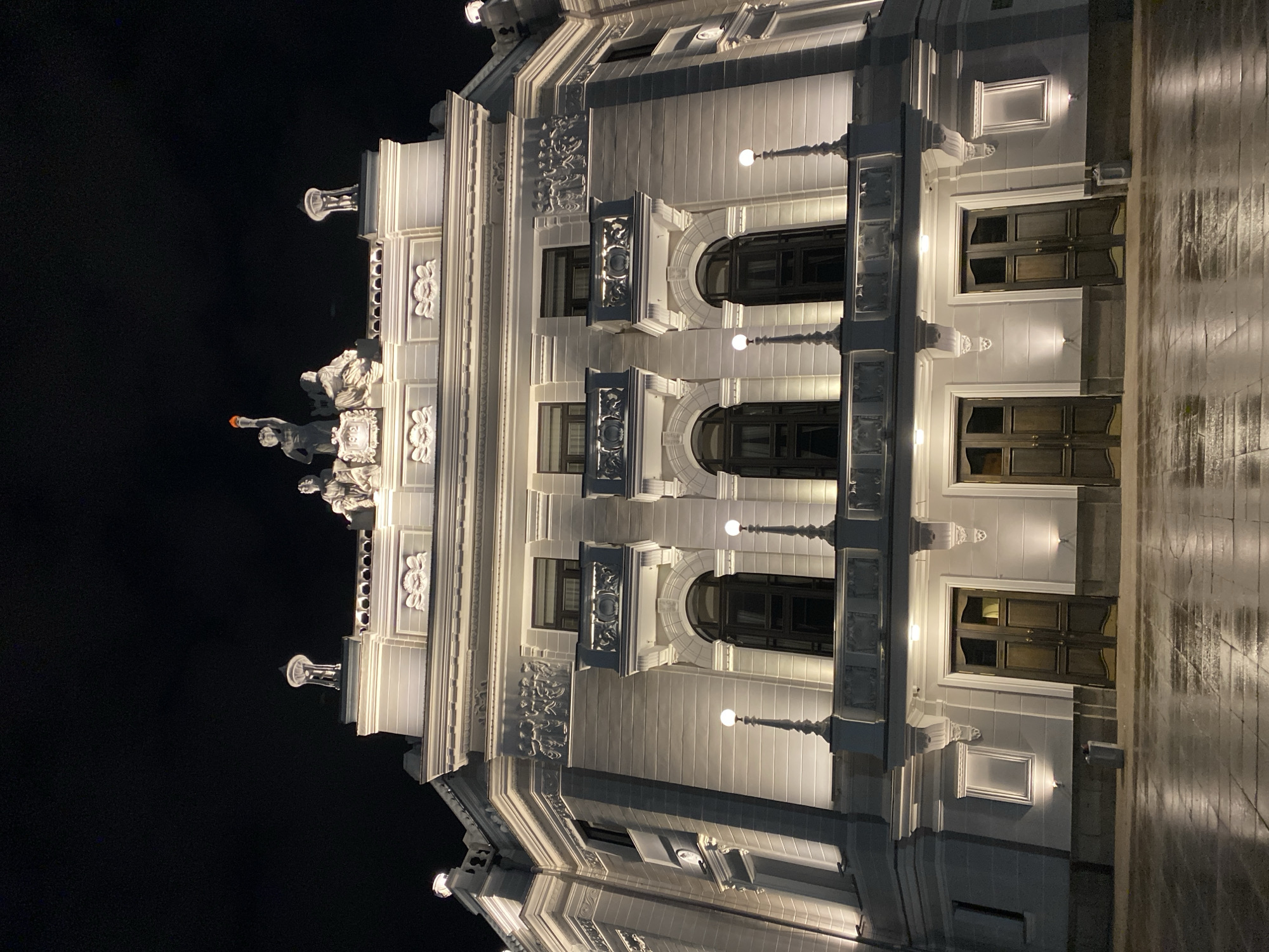A low-angle photograph depicts a portion of the white, illuminated facade of a building in Yekaterinburg, Russia, at night. The building has ornate architectural detailing, including decorative reliefs, small balconies, and a sculptural group at the top. Windows on multiple floors are visible, illuminated from within. Streetlights illuminate the facade, casting light and shadow. The sky is dark, and the wet sidewalk reflects the building's lights.