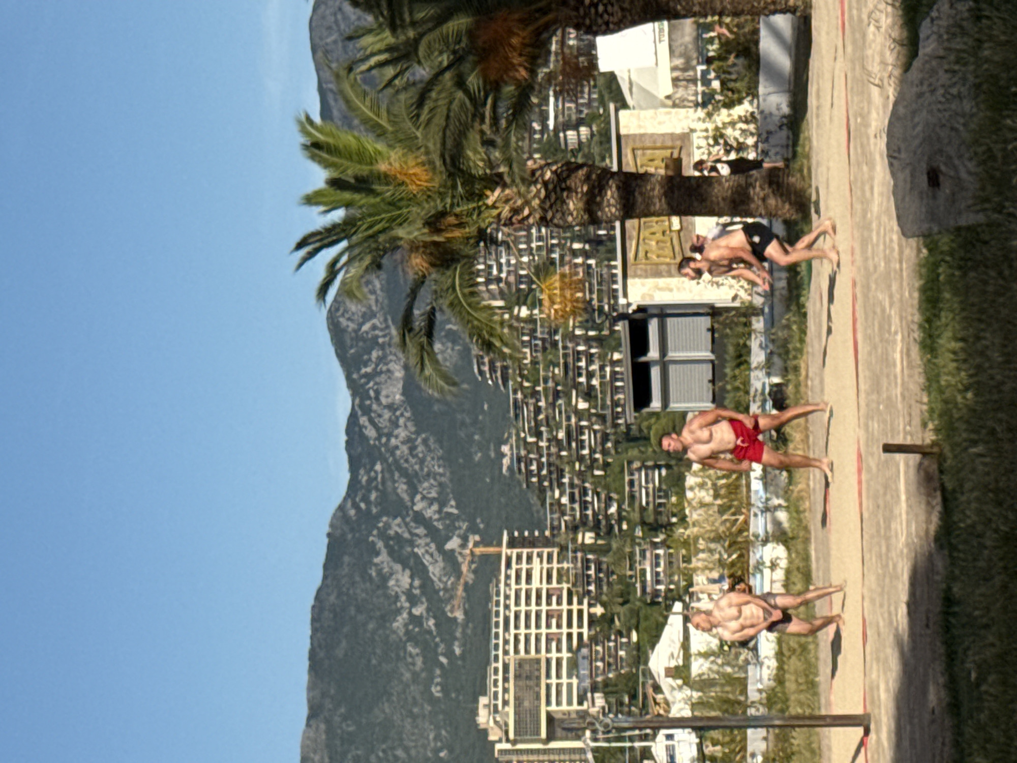 The image shows a beach volleyball game in progress. Four men are visible on a sand court. Two players are on one side of the net, while two are on the other. The man on the far left is wearing grey shorts, and the man to his right is wearing red shorts. On the other side of the net, one man is wearing black shorts, and another is also wearing black shorts. The background features palm trees, a multi-story building with a distinctive architectural style, and a mountainous landscape. The sky is clear and blue. The location is identified as Budva, Montenegro, suggesting a coastal Mediterranean setting.
