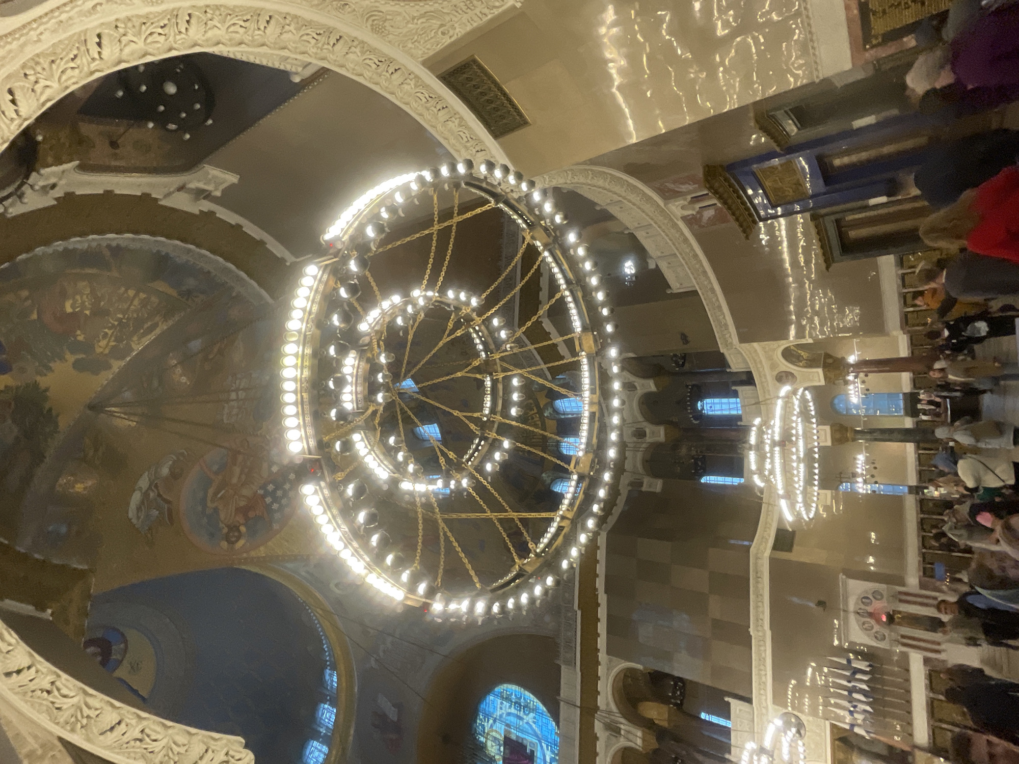 The image depicts the interior of the St. Nicholas Naval Cathedral in Kronstadt, Russia. The photograph is taken from a low angle, looking upwards towards the ornate dome and a large, multi-tiered chandelier. The chandelier is a prominent, golden structure adorned with numerous lights, casting a warm glow throughout the vast space. Above, the dome features painted murals depicting religious scenes.
Several other, smaller chandeliers are visible in the background, as are arched windows with blue stained glass. The walls of the cathedral are decorated with architectural embellishments and what appear to be plaques or memorials. The lower portion of the image shows a crowd of people, suggesting it is a public gathering. They are diverse in age and attire, and their interactions are not clearly discernible. The overall impression is one of grandeur and historical significance.