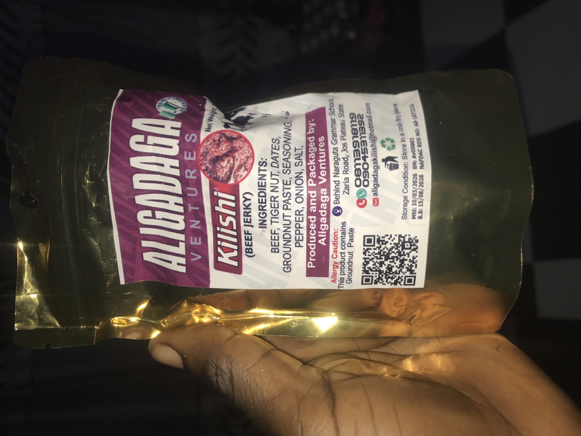 A close-up shot features a person's hand holding a golden, reflective stand-up pouch of a food product indoors. The package contains "Kilishi (BEEF JERKY)" by "ALIGADAGA VENTURES".
The product label, primarily purple and white, prominently displays the brand and product name. Key information visible on the label includes:
- **Ingredients:** BEEF, TIGER NUT, DATES, GROUNDNUT PASTE, SEASONING, PEPPER, ONION, SALT.
- **Producer and Packager:** Aligadaga Ventures, with an address specified as "Behind Naraguta Grammar School, Zaria Road, Jos Plateau State," correlating with the Rafin Par, Nigeria context.
- **Contact Details:** Phone numbers "08113919119" and "09045311392", and email "aligadagakilishi@hotmail.com".
- **Allergy Caution:** "This product contains Groundnut Paste".
- **Storage Condition:** "Store in a cool dry place".
- **Dates and Identifiers:** Manufactured date "10/03/2026", Best Before date "15/08/2026", Batch Number "Av00001", and NAFDAC Registration Number "A8-1072111".
- A QR code and recycling symbols are also present. The phrase "Net Weight" is partially visible.
The hand, with dark skin, supports the lower part of the pouch. The background is dark and featureless, indicating an indoor environment. The activity appears to be the presentation or display of the packaged food item, with clear emphasis on its details.