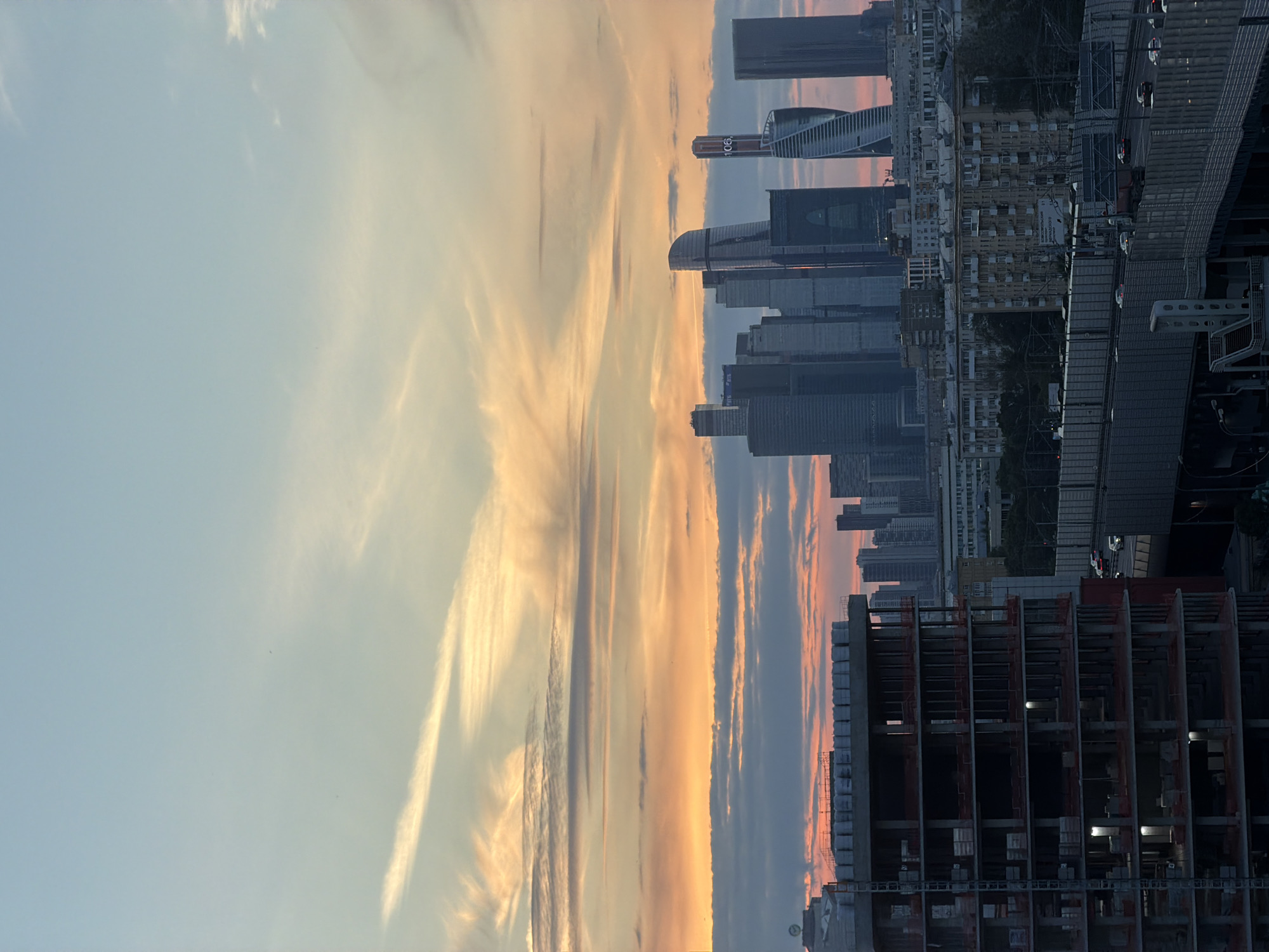 The image captures a skyline of Moscow, Russia, during sunset. In the foreground, a multi-story building under construction dominates the left side of the frame, with exposed concrete floors and scaffolding. To the right, a cluster of modern skyscrapers, including the Moscow International Business Center (MIBC), stands tall. The sky is painted with hues of orange, pink, and blue from the setting sun, with wispy clouds stretched across it. Below the skyscrapers, there are mid-rise residential buildings and a highway with cars visible. The scene appears to be a blend of ongoing development and established urban infrastructure.