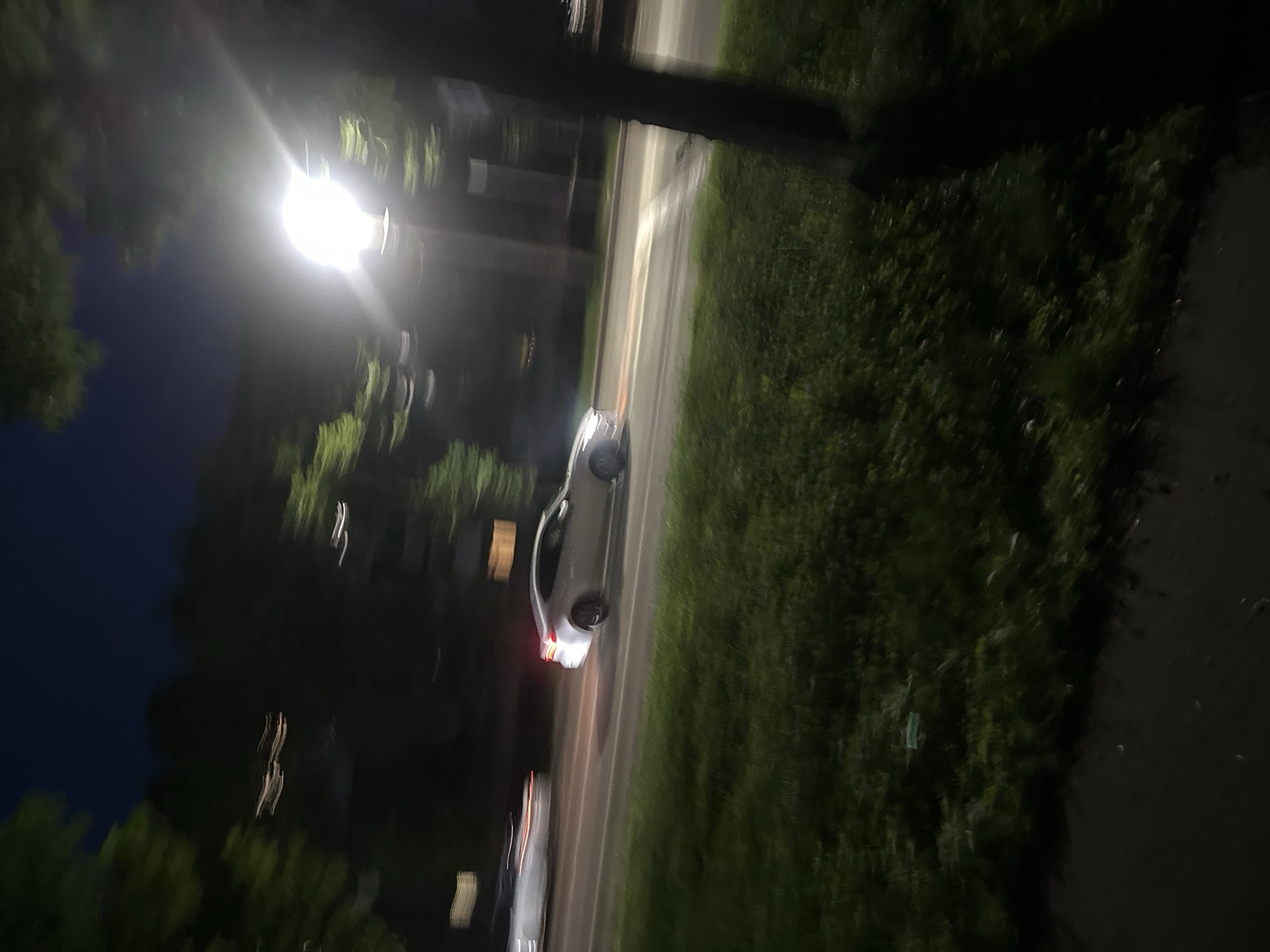 This media file depicts a nighttime scene, likely in Birobidzhan, Russia. The primary subject is a white car, possibly a sedan, driving or parked on a street. The image is characterized by motion blur, suggesting a long exposure or movement during capture, which creates streaking effects from lights and objects.
The scene is illuminated by a bright streetlight, casting a strong glow and creating lens flare. To the right of the street, there is a grassy area, and further to the right, a darker pavement or pathway. Trees with dark foliage are visible on the left side of the image, and a dark, possibly wooden pole, possibly a utility pole or part of a structure, is in the foreground, partially obscuring the view. The sky on the far left is dark blue, indicating nighttime. The motion blur of the car's taillights suggests it is either moving or has its brake lights illuminated. Another vehicle, lighter in color, is partially visible further down the road, also blurred.
The overall impression is one of a quiet urban or suburban street at night, with limited activity. There is no visible text in the image. The atmosphere is somewhat abstract due to the motion blur, focusing on light, shadow, and movement. The time of day is clearly night, and the weather appears clear, given the visibility of the stars or dark sky.