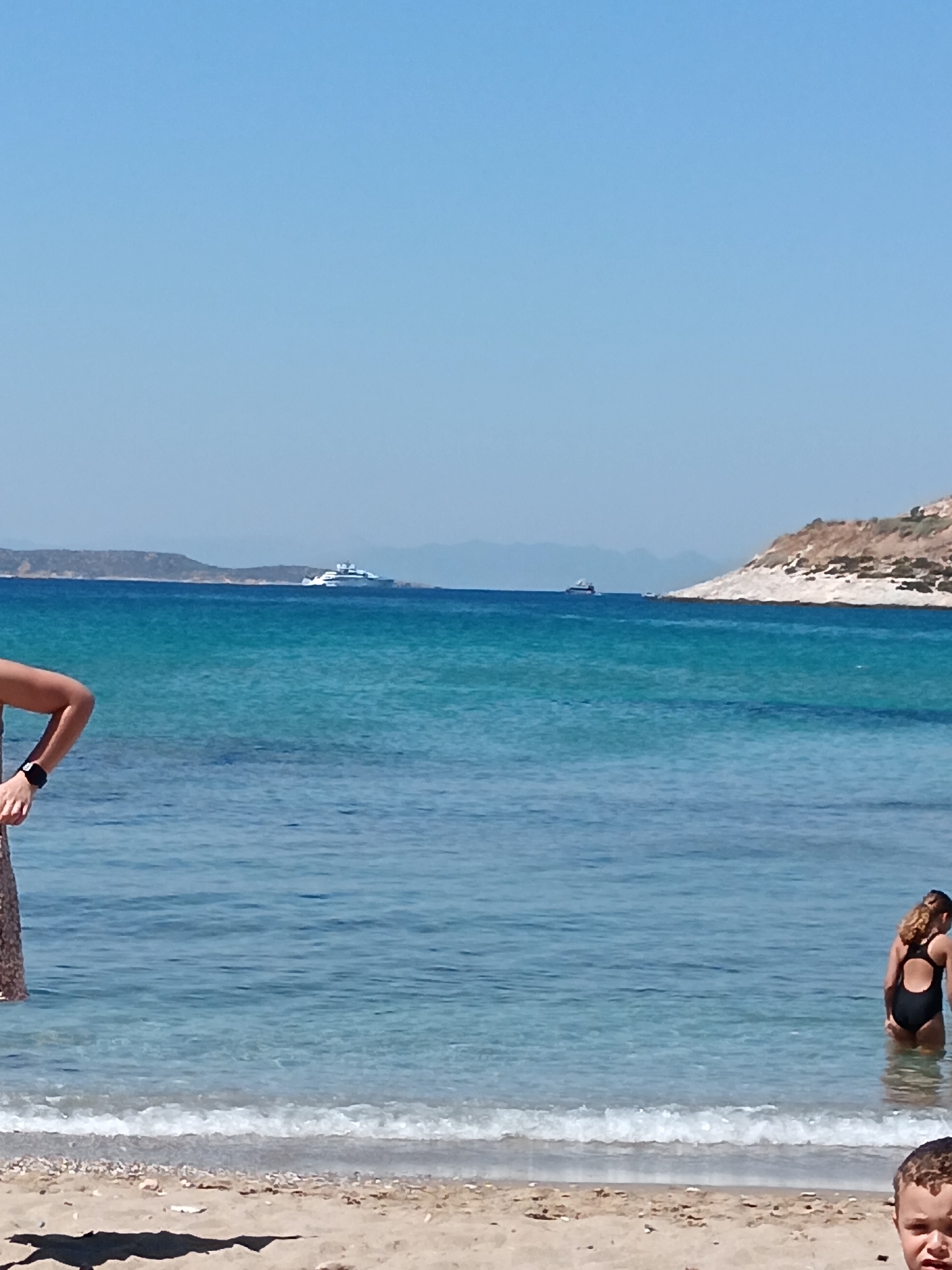 A wide-angle shot shows a serene beach scene under a clear blue sky. The turquoise waters of the Aegean Sea stretch out to the horizon, meeting a distant landmass. Two yachts are visible on the water, one larger and white, the other smaller and darker. A rugged, treeless island rises from the sea on the right side of the frame, its dry, rocky slopes contrasting with the vibrant blue water.
In the foreground, the sandy beach is sparsely littered with small pebbles and dried seaweed. The tide is gently lapping at the shore, creating a soft, white foam. On the left, a person's arm and part of their patterned skirt are visible, suggesting they are standing in the shallow water. On the right, a young girl in a black swimsuit stands in the water, her curly hair catching the sunlight. Slightly lower down and to the right of the frame, the face of a young boy with curly hair is visible, looking directly at the camera. The overall atmosphere is peaceful and summery, indicative of a warm, sunny day at a Greek beach, likely Agia Marina. There is no visible text in the image.
