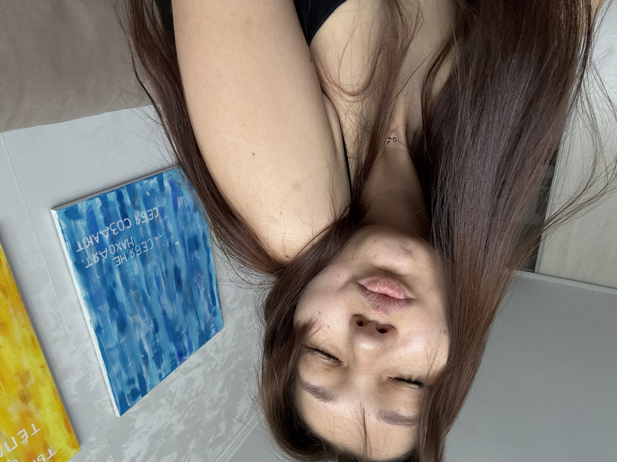 A person is lying down with their head tilted back, eyes closed and lips pursed as if blowing a kiss. Their long, brown hair falls around their face and shoulders. They are wearing a black tank top. Beside them is a small, rectangular canvas painted with vertical strokes of various shades of blue. Another canvas, yellow with some text, is partially visible to the left. The background appears to be a wall and a textured surface, possibly a bed or floor. The scene is likely indoors, given the lighting. The visible text on the blue canvas reads, "СЕБЯ НЕ НАХОДЯТ" (meaning "One doesn't find oneself").