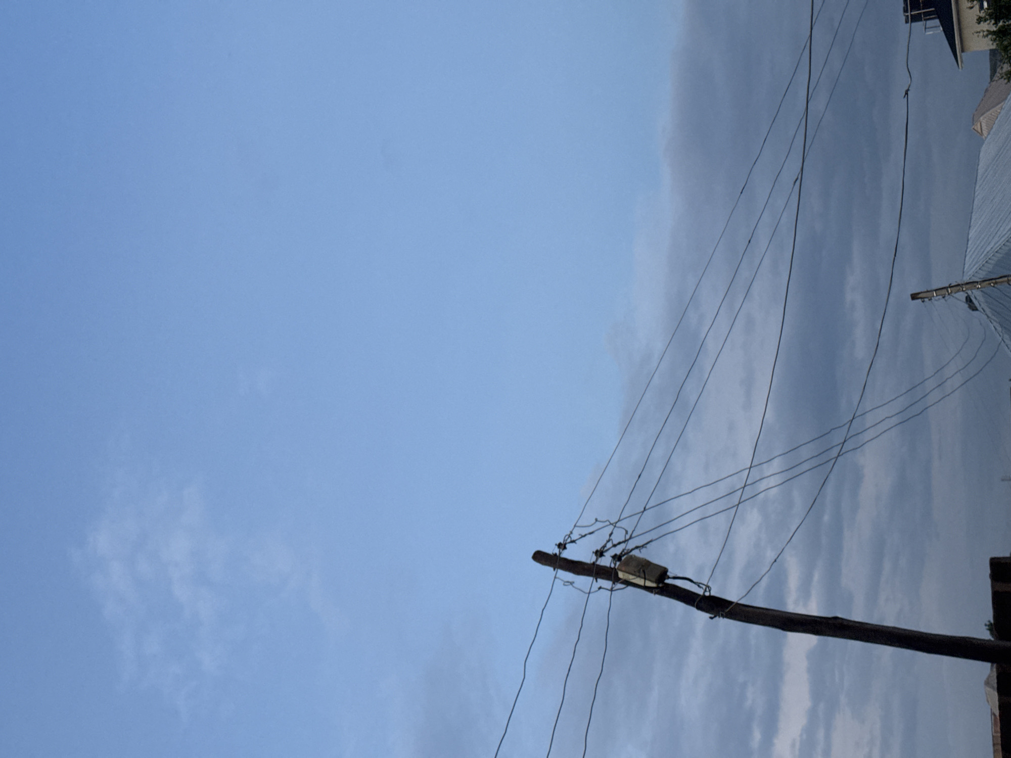 A low-angle, medium shot captures a wooden utility pole and power lines against a pale blue sky with scattered clouds in Unknown, Unknown. The pole is positioned on the left side of the frame, angled diagonally upwards, and is adorned with a gray electrical box and numerous wires. The wires extend across the frame, creating a network against the sky and partially obscuring some of the cloud cover. In the lower portion of the image, parts of rooftops are visible, suggesting a residential or urban environment. The lighting indicates it may be early morning or late afternoon.