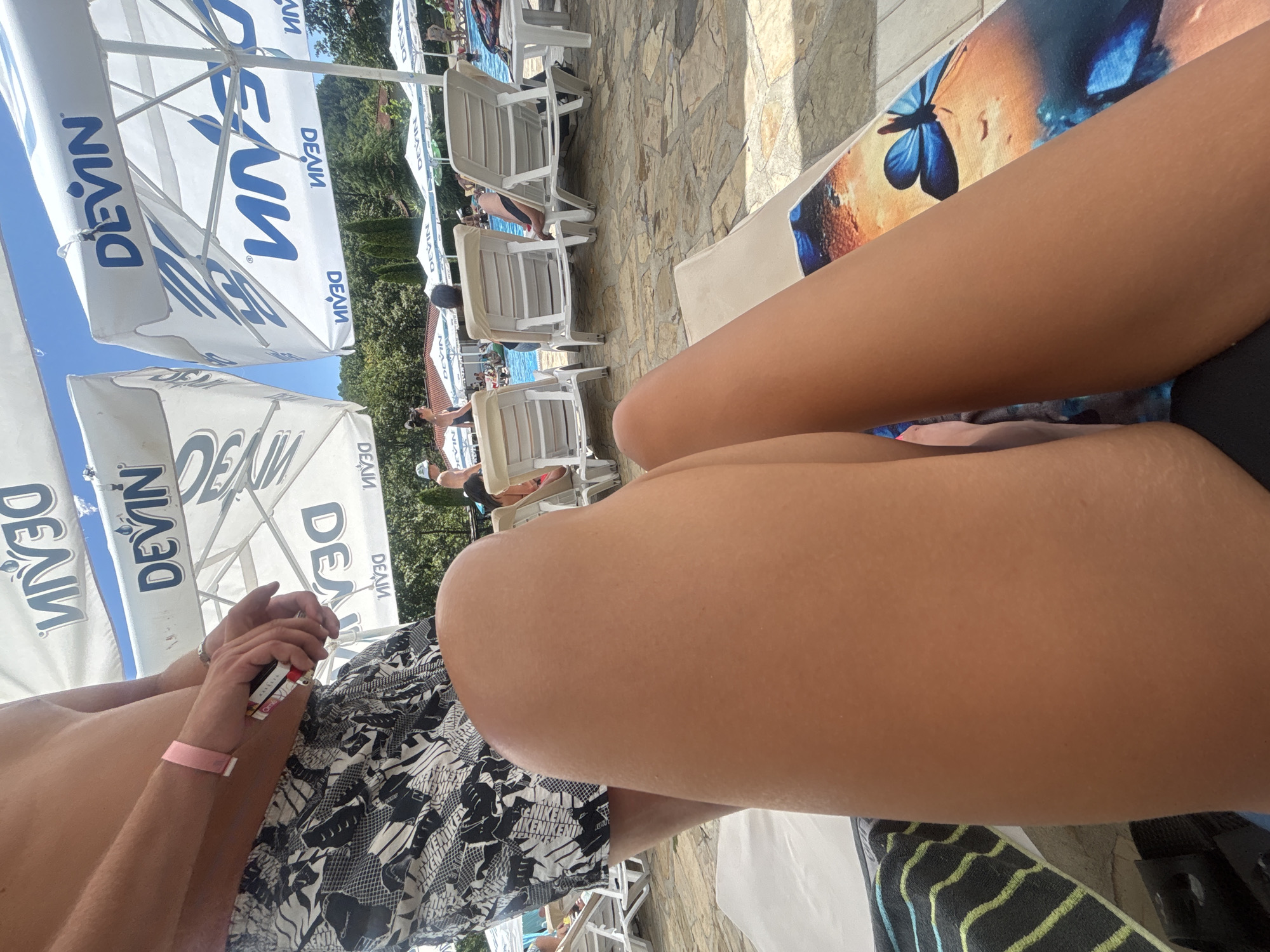 This image shows a person's legs and torso, lounging on a sunbed by a pool. The subject is wearing patterned swim trunks and a pink wristband. In their hand, they are holding a cigarette lighter. The scene is outdoors, with several white beach umbrellas visible in the background, some bearing the blue text "DEVIN". There are also lounge chairs, a pool, and trees in the background, suggesting a sunny day at a resort or pool area. The overall impression is one of relaxation and leisure.