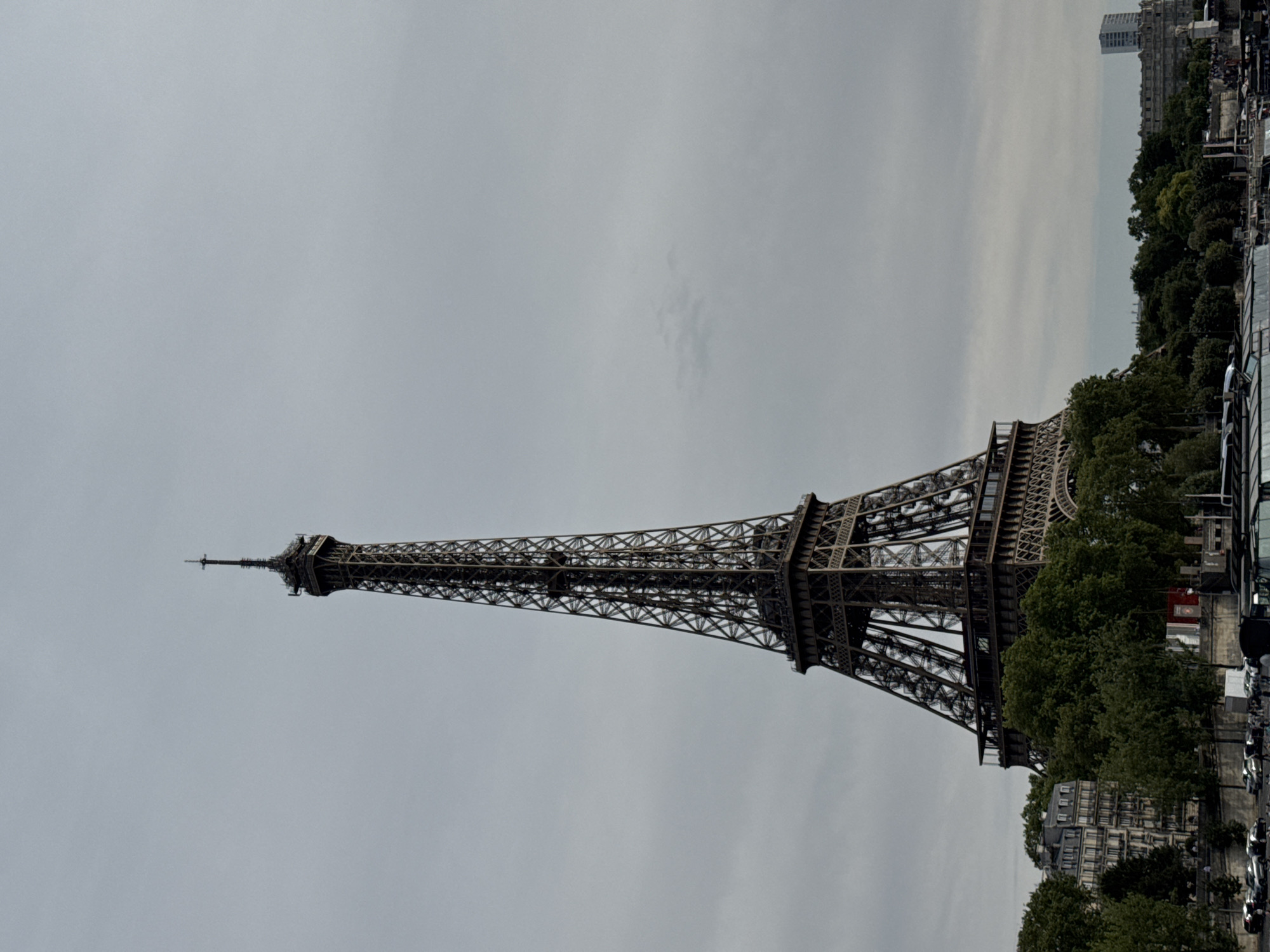 The image shows the Eiffel Tower in Paris, France, on a cloudy day. The tower is viewed from below, with the sky taking up most of the frame. Trees and some buildings are visible at the base of the tower, and a body of water, likely the Seine River, is in the foreground. Several boats are docked along the river. Cars are parked on the side of the road near the trees and buildings. A few people are visible in the distance near the water and along the bank. The overall atmosphere is somewhat overcast and muted.