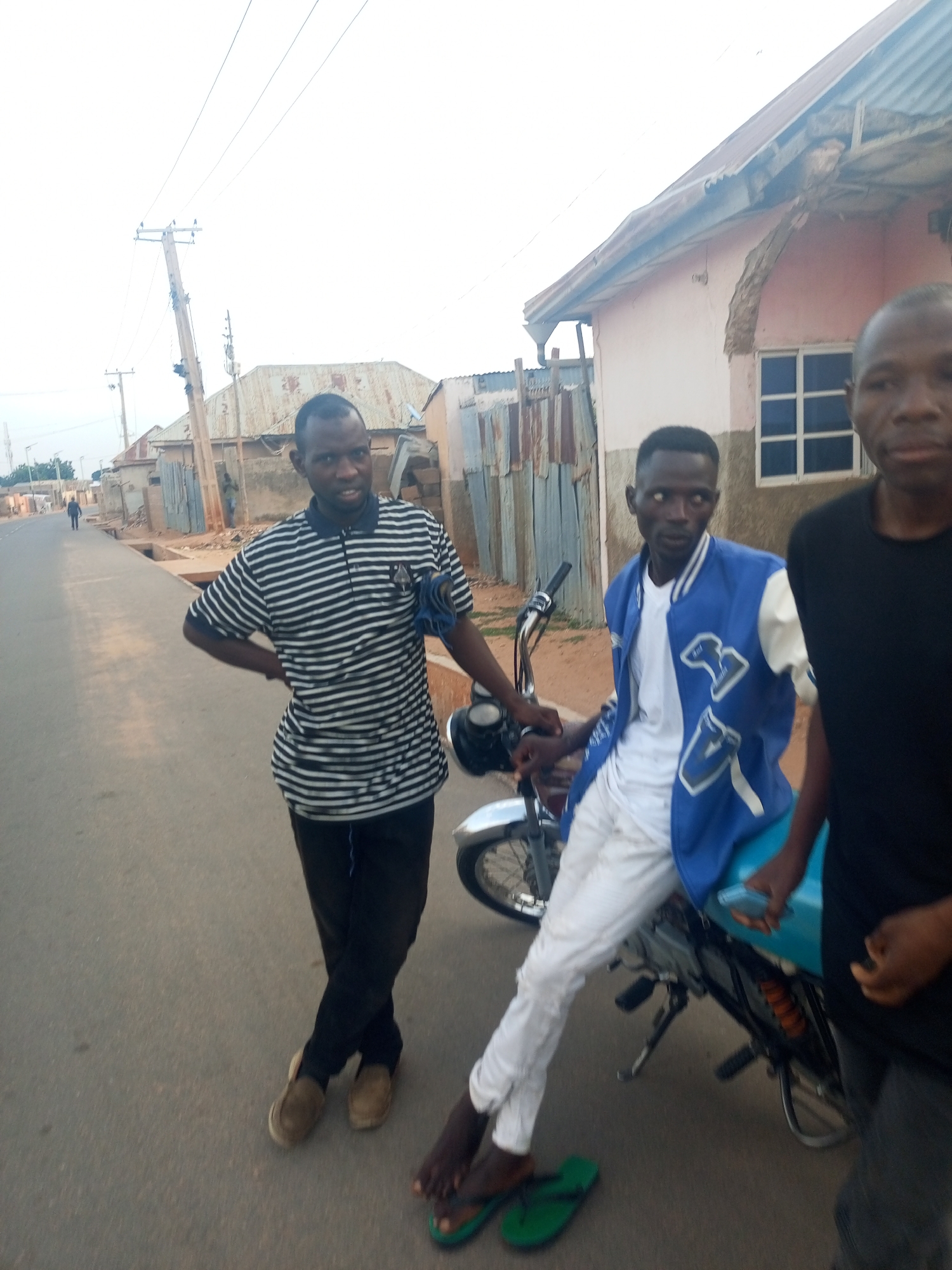 Three males stand on a paved road alongside a motorcycle in what appears to be a city neighborhood. The male on the left wears a striped black and white polo shirt with black pants and brown shoes, leaning against the motorcycle. The central male is seated on the motorcycle, wearing a white t-shirt, blue jacket, and white pants, with green flip-flops near his feet. The male on the right is partially visible, wearing a black t-shirt. The environment includes buildings, some with corrugated metal siding, and overhead power lines. A lone individual walks in the distance on the road. The general location is in the city of Unknown, Unknown.