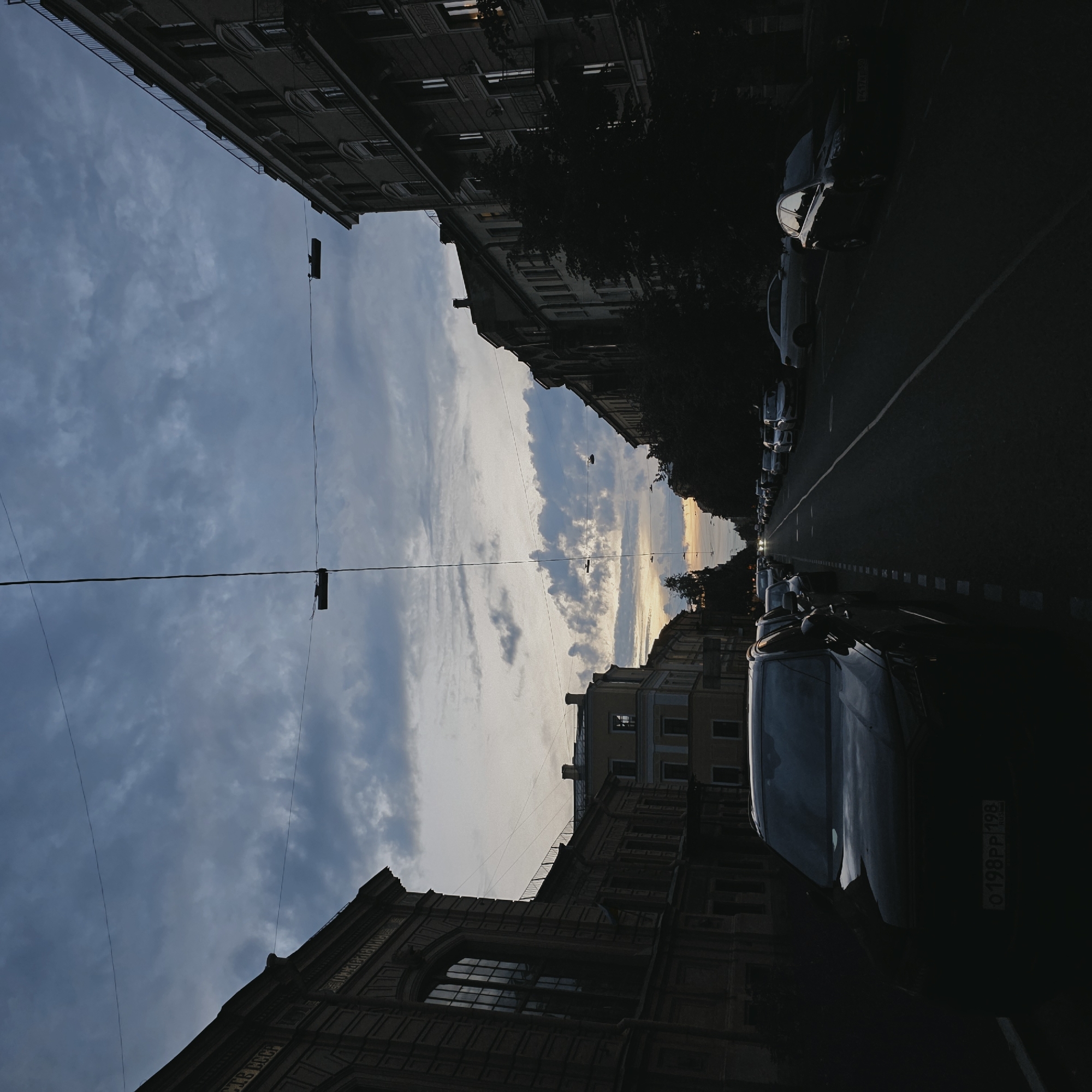The image depicts a street scene in Saint Petersburg, Russia, during twilight. The perspective is from a street-level viewpoint, looking down a relatively straight road lined with multistory buildings on both sides. The sky is overcast with dramatic clouds, tinged with the soft hues of sunset. Overhead, a network of wires, possibly for streetlights or trams, crisscrosses the sky, adding a linear element to the composition.
On the left side of the street, a dark-colored SUV is parked prominently in the foreground, its front end slightly angled towards the viewer. Its license plate is partially visible, showing the characters "0198PP 198" and the word "RUS". Beyond this vehicle, other cars are parked along the curb, receding into the distance.
The buildings flanking the street are typical of St. Petersburg architecture, with ornate facades and numerous windows. The building on the left is a darker color, possibly brick, with decorative molding around its windows and along the roofline. The buildings on the right appear lighter in color, with balconies on some floors.
The street itself is paved and appears to be a two-way road with a clear lane marking. Parked cars line both sides of the street, creating a sense of urban density. Trees are visible on the right side, their dark foliage contrasting with the sky and buildings.
In the distance, the road appears to curve slightly, and the faint glow of streetlights or car headlights can be seen, indicating the deepening twilight. The overall atmosphere is quiet and slightly moody, characteristic of an urban environment at the end of the day.
