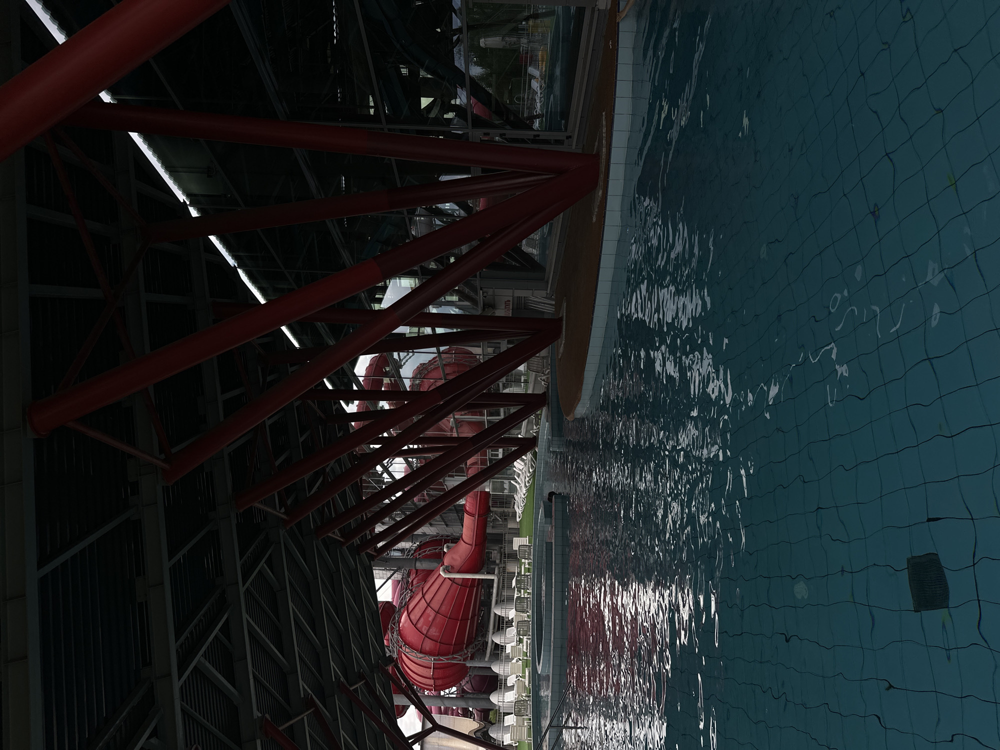 The image depicts the interior of a water park. In the foreground, a swimming pool with tiled bottom and clear blue water is visible. The water's surface reflects the overhead structure and artificial lighting. In the mid-ground, a wooden deck area borders the pool, and beyond that, white lounge chairs and some spiral structures are arranged on a green surface. Dominating the background are large, bright red water slides, curving and twisting within the enclosed structure. The entire scene is supported by a complex network of dark metal beams and red, angular support columns. The environment appears to be a climate-controlled indoor facility. The location is specified as Minsk, Belarus, likely referring to a specific water park within the city, such as "Aqua Park Lebyazhy". There are no people clearly visible in the image, nor any specific actions or interactions taking place. The scene is static and empty, suggesting it was captured during off-hours or without visitors present.