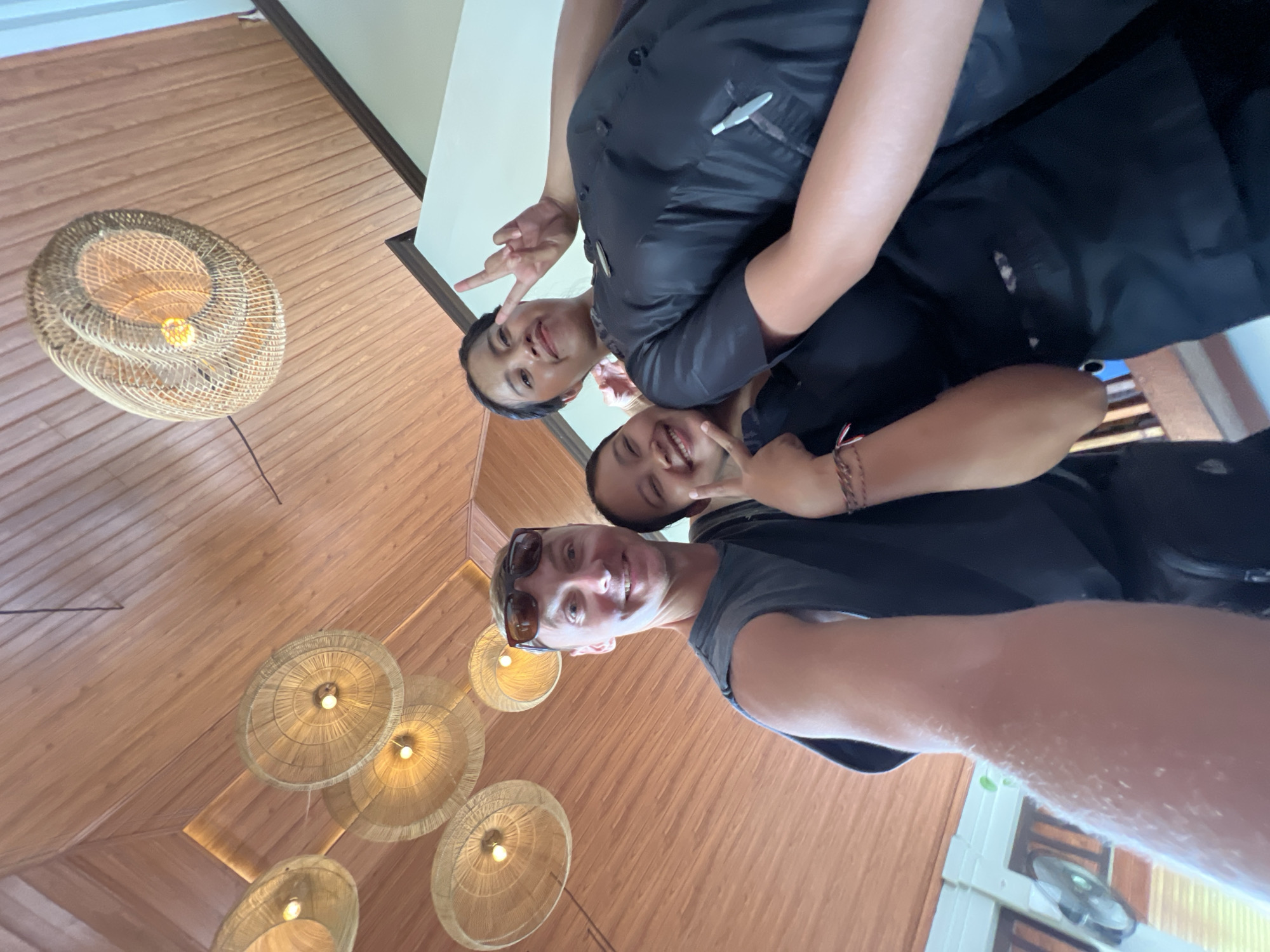 Three individuals are closely grouped together in a selfie-style photograph taken from a low angle. The background consists of wooden walls and numerous wicker pendant lights. The location appears to be indoors, possibly a hotel or restaurant within the city of Kuta Selatan, Indonesia. The lighting is warm and artificial.
The person closest to the camera is a fair-skinned male wearing sunglasses and a gray t-shirt. He is smiling and holding the camera. Behind him is another individual with darker skin, also smiling. A third individual is positioned behind and slightly to the side, making a peace sign with their fingers. The clothing worn by the two individuals in the background is predominantly dark or black.