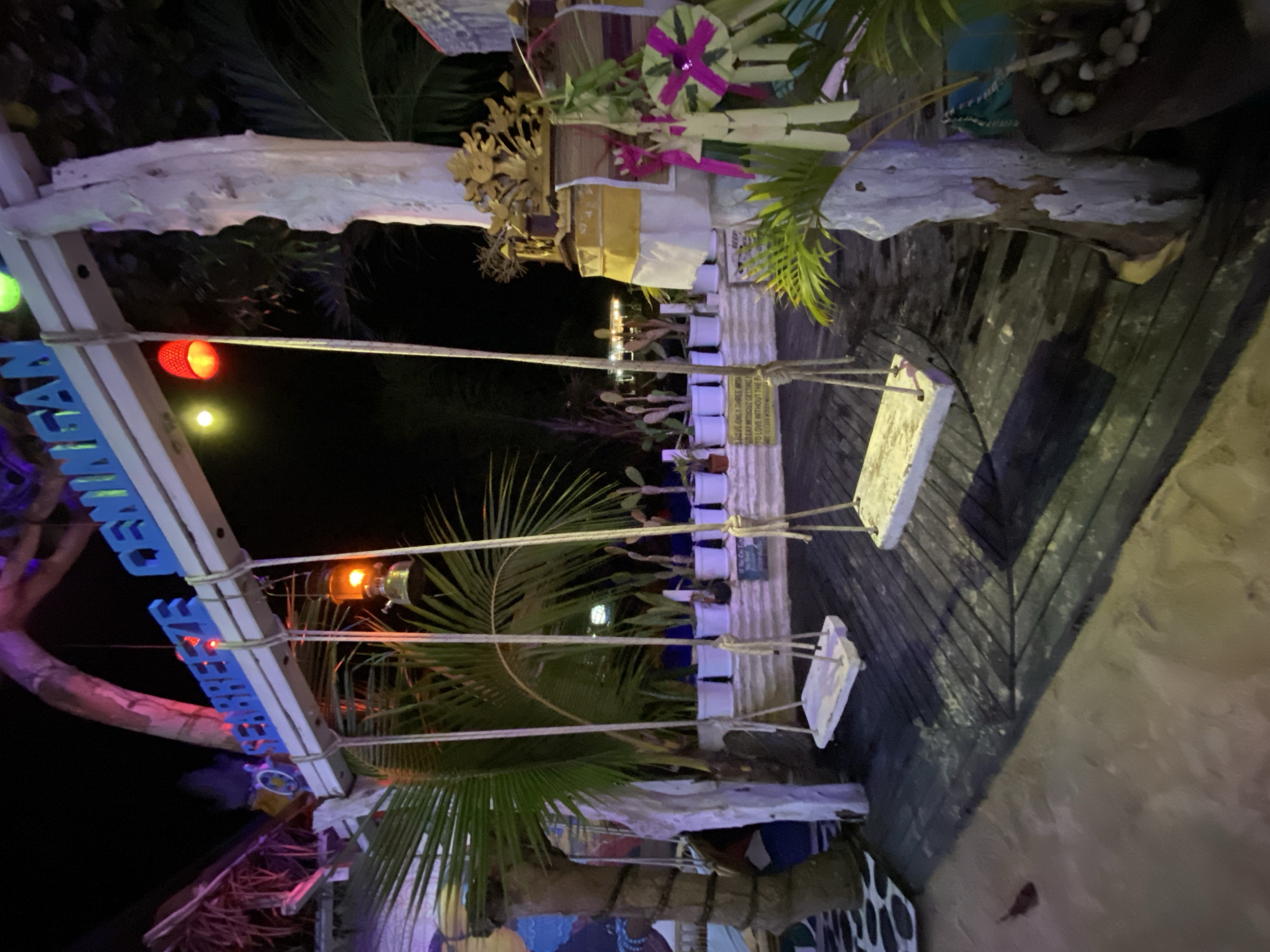 This media file captures a nighttime scene at a beachside establishment, likely a bar or restaurant, in Nusa Penida, Indonesia. The setting is a wooden deck bordering a sandy beach, illuminated by soft artificial lights.
The primary subjects are two swings made of white planks suspended by thick ropes from a rustic wooden structure. These swings are positioned over the wooden deck. There are several other objects visible, including potted plants in white containers, a decorative arrangement of palm leaves and fabric, and a weathered piece of driftwood. A blue sign with white lettering spelling out "SEMINYAK" is attached to a wooden post on the left side of the frame. Small lights, including a red one and an orange one, are attached to the structure supporting the swings and the sign.
The scene appears to be set at night, as indicated by the dark sky and the artificial lighting. The weather seems calm and pleasant, suitable for an outdoor evening at the beach. The overall atmosphere suggests a relaxed and inviting ambiance, typical of a tropical vacation spot. There is no activity or event clearly depicted; it appears to be a static shot of the location.
Notable details include the tropical foliage, such as palm fronds, contributing to the island setting. The weathered wood and rustic decor suggest a casual, perhaps bohemian, style. The text "SEMINYAK" is clearly visible on the blue sign, which is a well-known tourist area in Bali, but its presence here in Nusa Penida might indicate a themed establishment or a direct reference to a popular location.