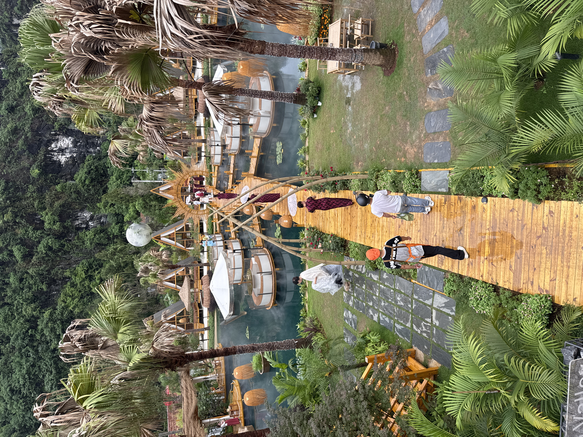 This image shows an aerial view of a tourist attraction located in Guilin, China. The scene includes a network of wooden walkways and floating platforms on a body of water. Lush green foliage and palm trees surround the area. Several people are walking on the walkways, some dressed in traditional clothing. Bamboo structures and decorative lanterns enhance the setting. One person is wearing a bright orange hat and harness. The setting appears to be a designed outdoor leisure or entertainment space.