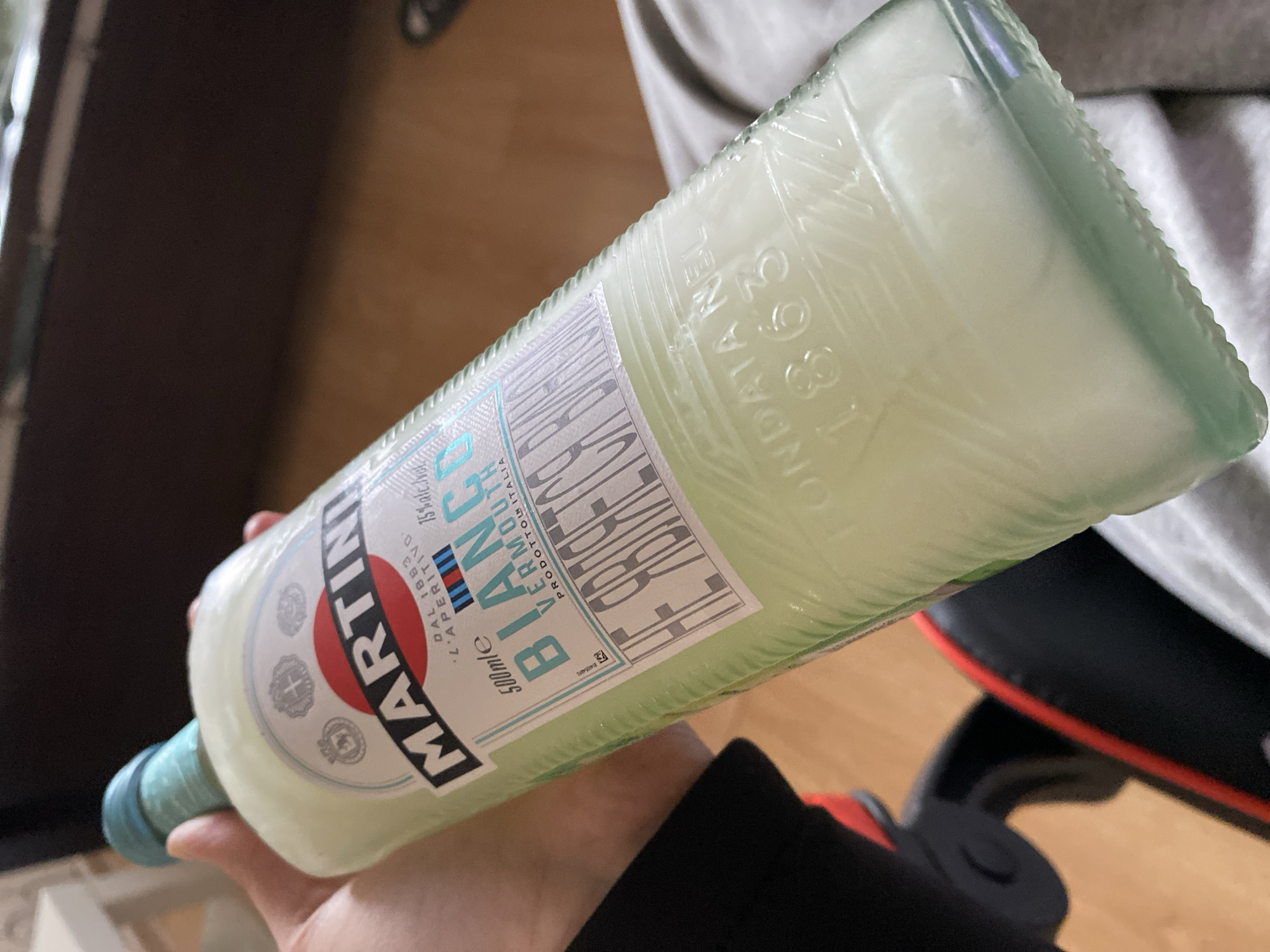 A person is holding a bottle of Martini Bianco vermouth. The bottle is green glass and appears to be partially full with a pale, yellowish liquid. The label on the bottle indicates "MARTINI" in bold letters with the words "Bianco" and "Vermouth" below. The text "DAL 1883 L'APERITIVO" and "PRODOTTO IN ITALIA" are also visible. The bottle size is "500mle" with an alcohol content of "15% alc./vol.". The text "НЕ ЯВЛЯЕТСЯ ВИЛНО" is also present on the label. The bottle's texture and lettering on the upper part read "FONDATA NEL 1863". The environment appears to be indoors, with a blurred background of furniture or clothing. The location is not specified but the presence of Russian text on the label indicates it is available for purchase in Russia, but does not mean the image was taken in Izhevsk.
