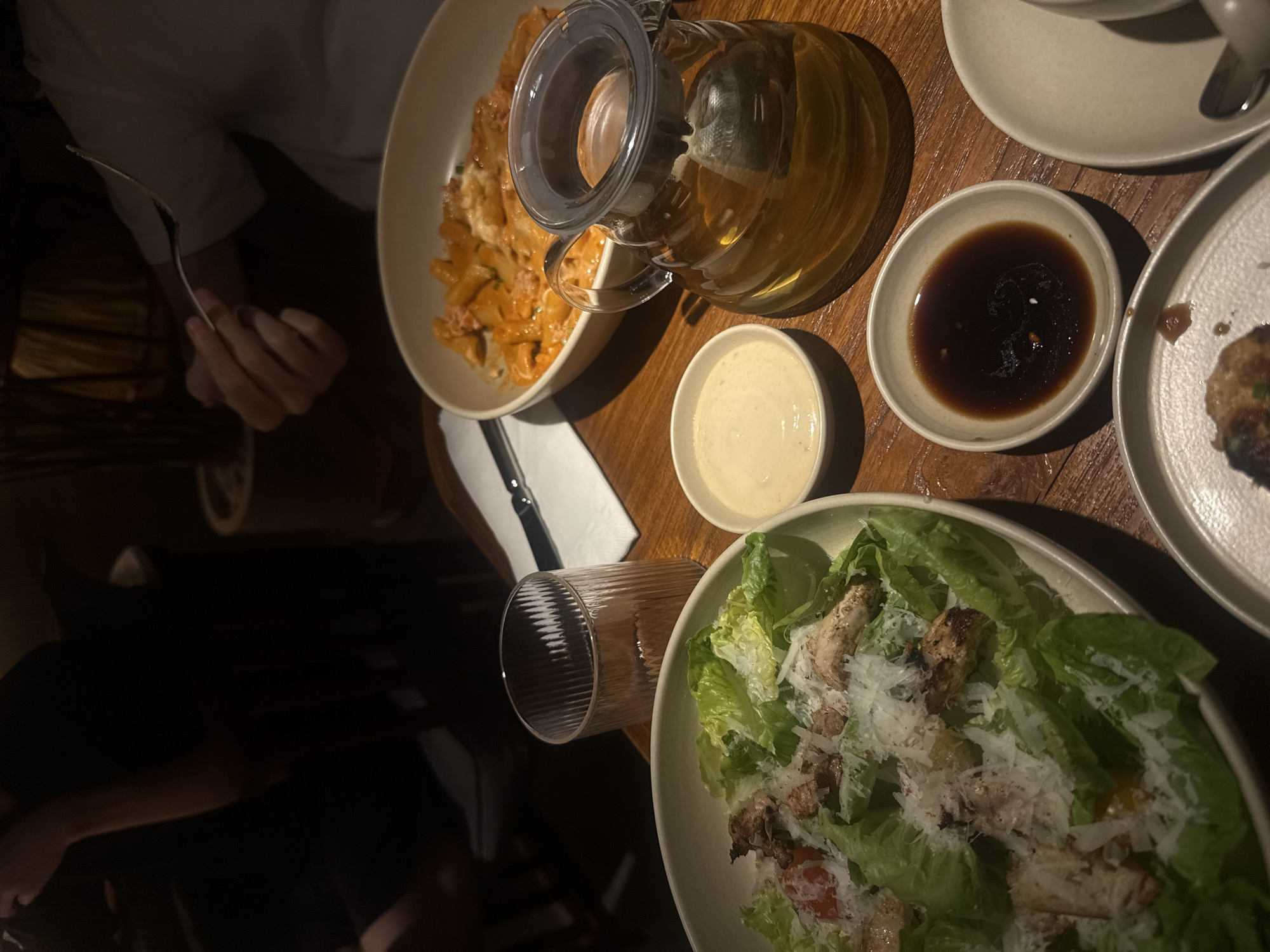 The image presents a top-down view of a dining table laden with food and drinks in a dimly lit indoor setting, likely a restaurant or a home during an evening meal. The table surface is a polished dark wood.
In the upper left, a white bowl holds a pasta dish, possibly penne, covered in a creamy, reddish sauce and topped with white grated cheese. Beside it, a clear glass teapot with a black lid contains a light amber beverage, likely tea, with a tea bag visible inside.
To the right of the teapot, a small, light-colored bowl holds a dark brown sauce. Below the teapot, another small bowl contains a pale, creamy white sauce. In the bottom right portion of the frame, a large white bowl features a fresh green salad, consistent with a Caesar salad, featuring crisp lettuce, pieces of grilled chicken, and a generous dusting of grated cheese. Some red components, possibly cherry tomatoes, are also visible in the salad.
A person's arm and upper torso, dressed in a light-colored shirt, are visible on the far left, with their hand holding a fork, poised to eat the pasta. Another person's arm in a dark shirt is subtly visible at the very bottom edge. A ribbed clear drinking glass stands empty near the salad, and a folded white napkin with a knife rests between the pasta bowl and the empty glass.
The overall atmosphere is warm and inviting due to the low lighting. No text is visible in the image.