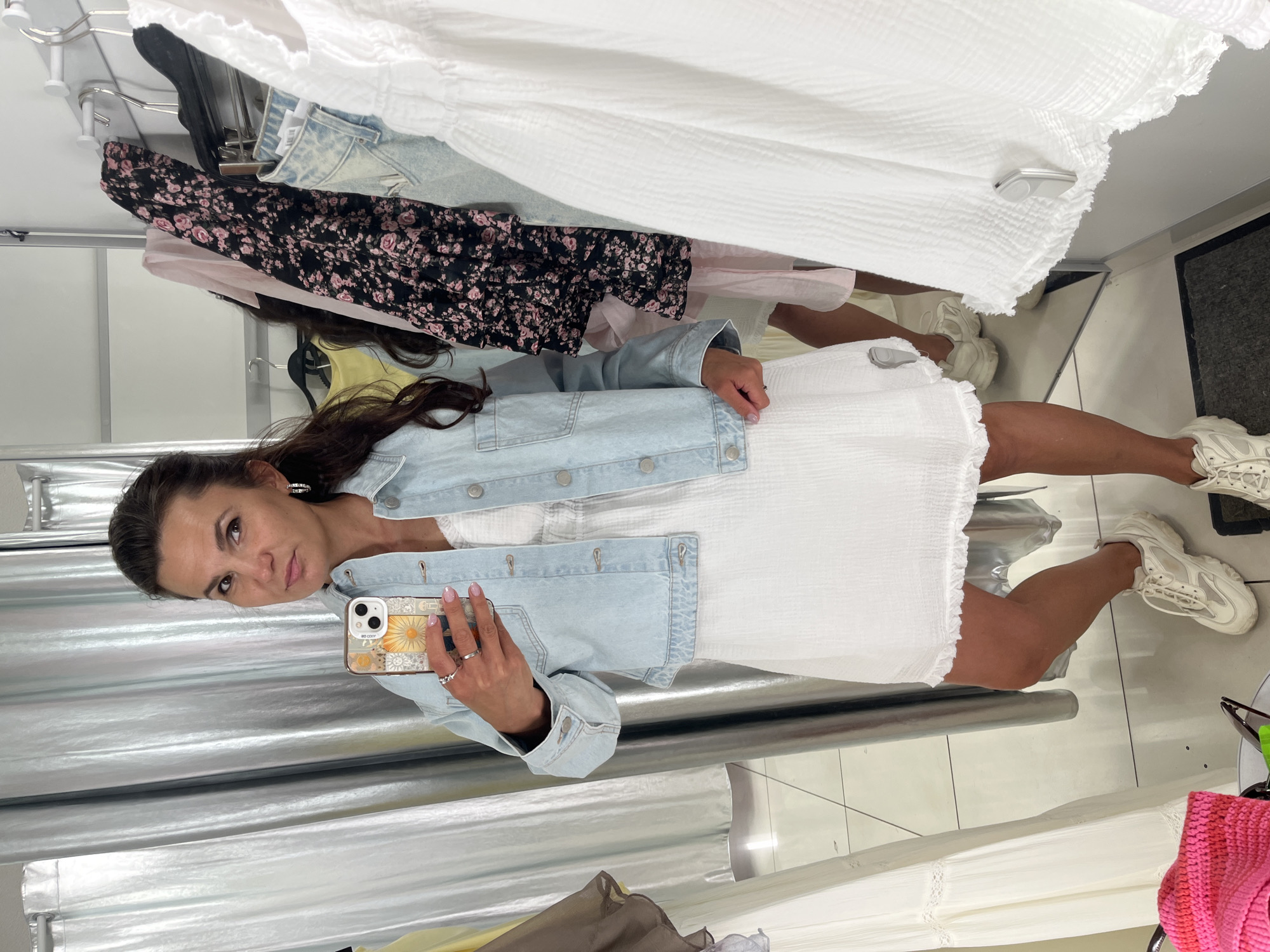 In a clothing store fitting room, a woman stands facing a mirror, taking a selfie with her phone. She is wearing a light blue denim jacket over a white dress and white sneakers. The fitting room is lined with racks of clothing and has silver, floor-to-ceiling curtains. The woman is holding her phone, which has a colorful case with a floral design, in her right hand. Her left hand is visible with rings on her fingers. In the background, various garments hang on racks, including a black floral dress and white dresses. The flooring appears to be tiled.