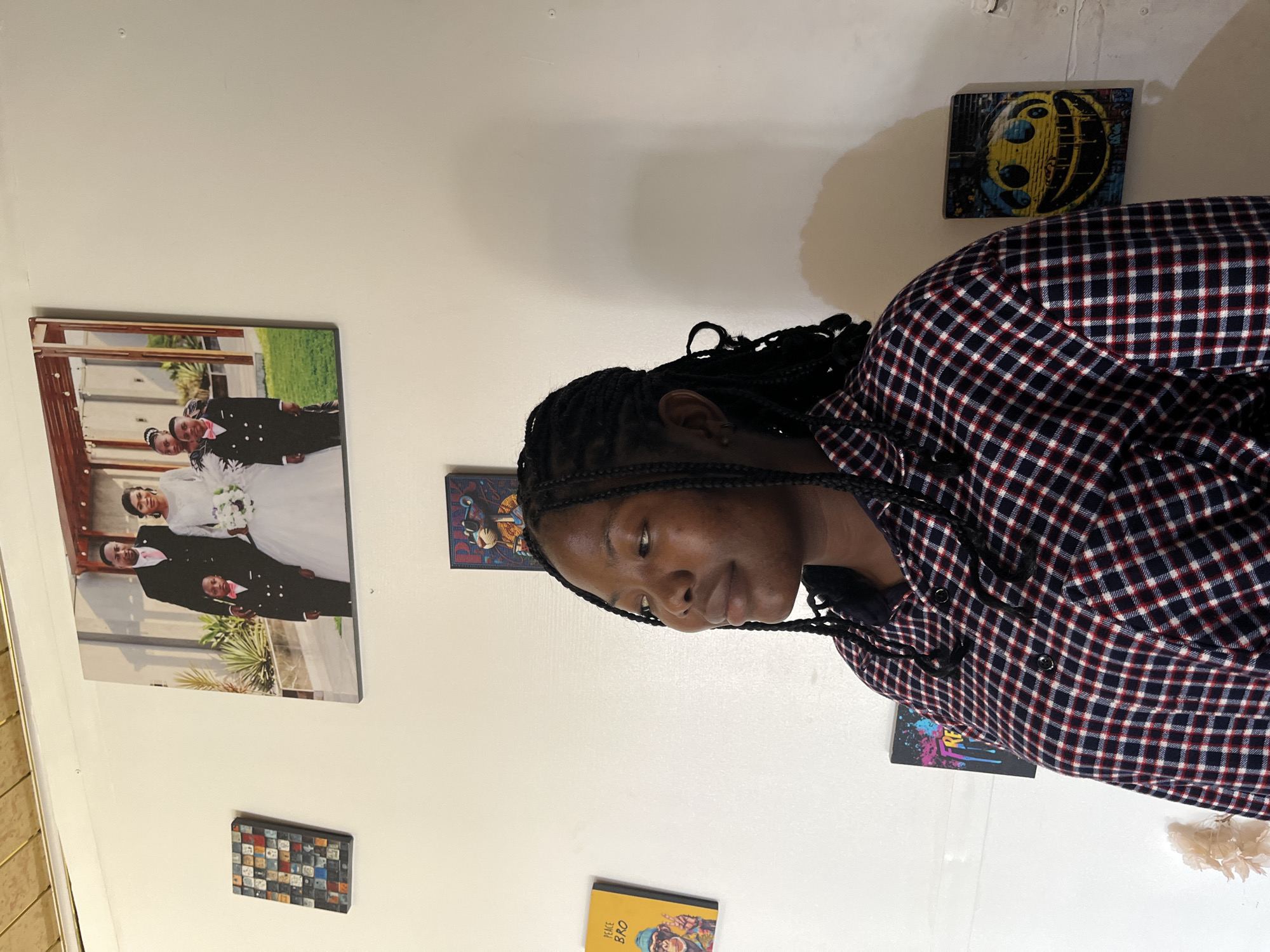 The image shows a young woman with braided hair standing in front of a white wall decorated with several framed artworks. She is wearing a plaid shirt. To her left, there are three small framed pictures on the wall. The top picture is a wedding photograph of a couple with two young boys. Below that, to the left of the woman, is a yellow framed picture with a monkey graphic and the text "PEACE BRO". To the right of the yellow picture, and above the woman's shoulder, is a small framed artwork depicting a bear wearing sunglasses. To the right of the woman, there is a small framed artwork featuring a dripping smiley face. The overall setting appears to be an indoor residential space, possibly a living room, with a warm lighting. The image is captured from a medium distance, focusing on the woman and the wall decorations. There is no information in the image to suggest a specific location in Jos, Nigeria.