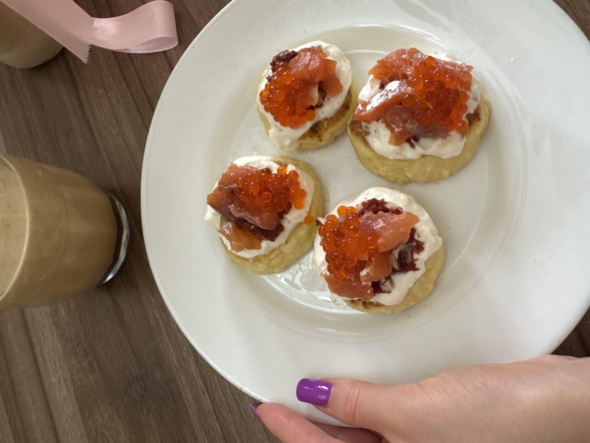 The image depicts a plate of four small, round appetizers, each topped with white cream, what appears to be smoked salmon, and orange roe. The plate is white and is held by a person's hand with purple nail polish. To the left of the plate is a glass of light brown liquid and a pink ribbon. The background is a wooden surface.