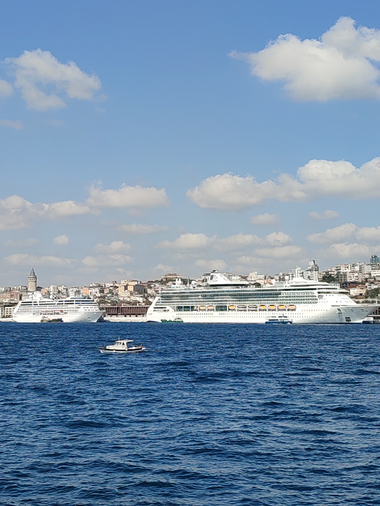 The image captures a wide view of the Bosphorus Strait in Istanbul, Turkey, with the Beyoğlu district visible in the background. Two large white cruise ships are docked, appearing majestic against the cityscape. To the left, the iconic Galata Tower stands tall, a historical landmark of Beyoğlu. The water is a deep blue, with ripples suggesting gentle movement. Several smaller boats are scattered across the strait, including one in the foreground that is small and white. The sky is a bright blue, dotted with fluffy white clouds. Buildings with a mix of architectural styles line the shore of Beyoğlu, creating a dense urban backdrop. The overall scene depicts a bustling harbor with a blend of modern and historical elements.