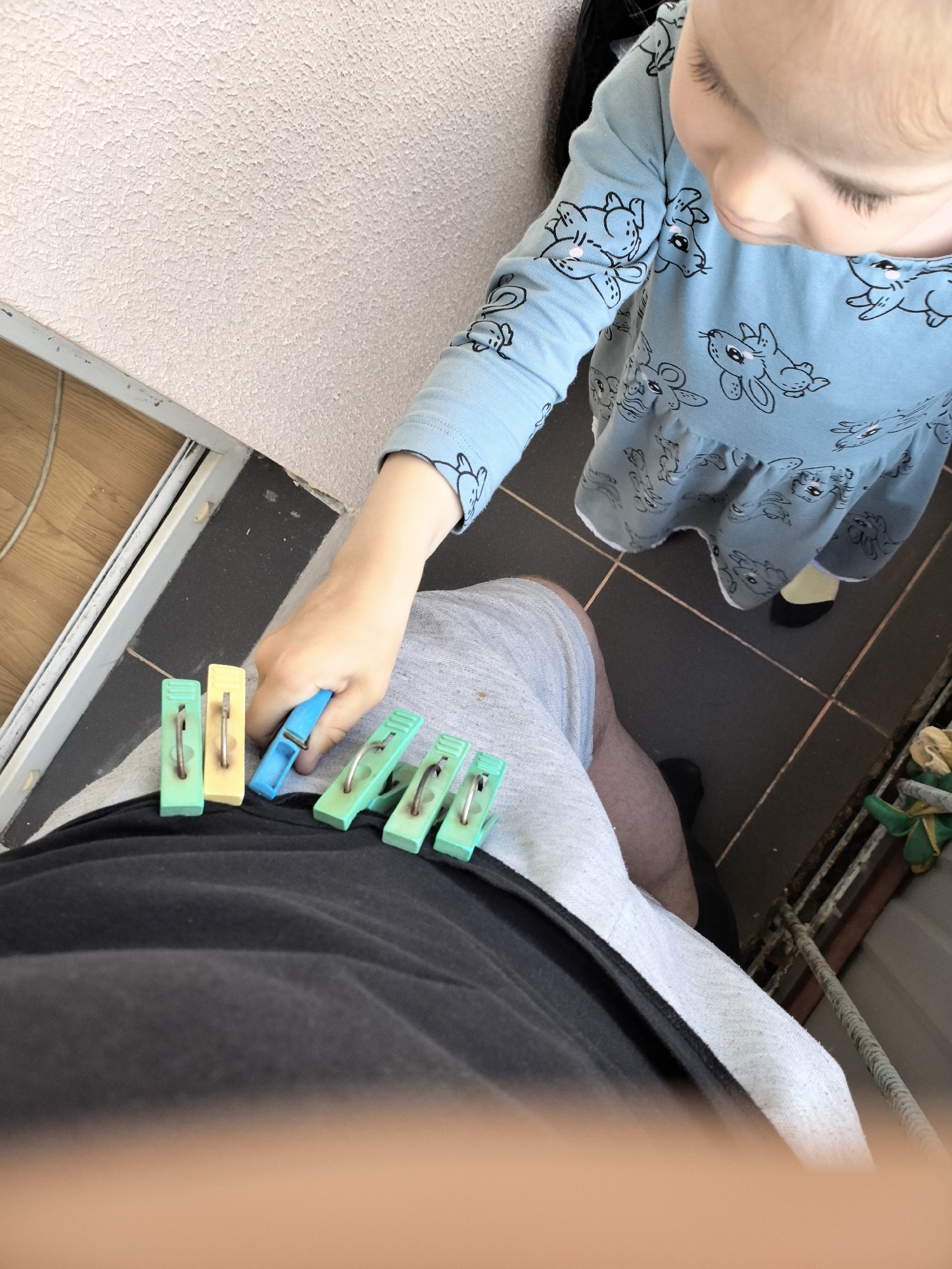 An overhead, close-up shot shows a toddler's hand holding a blue plastic clothespin while reaching for several other clothespins lined up on a black t-shirt. The clothespins are green and yellow. The toddler wears a light blue long-sleeved dress with a pattern of cartoon bunnies. They have fair skin and light hair. The toddler's face is partially visible, looking down at their hand. The scene appears to be outdoors, possibly on a balcony, with a textured pink wall visible in the upper left corner and dark grey tiled flooring. The image is taken from the perspective of someone lying down.