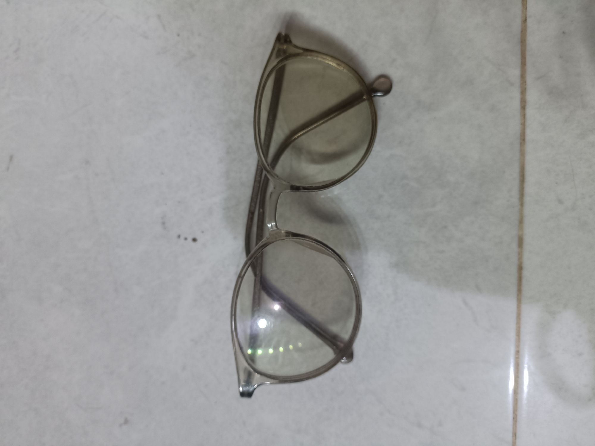 A pair of clear-lensed glasses with transparent frames lies on a light-colored tiled surface. The glasses are positioned vertically in the center of the frame, with the lenses facing upwards and slightly tilted. The right lens reflects several bright, greenish-yellow lights. The tiled surface appears to be part of an indoor environment, possibly a floor, and shows some subtle variations in color and texture. There are no people or any other objects visible in the image, apart from the glasses and the tiled background.