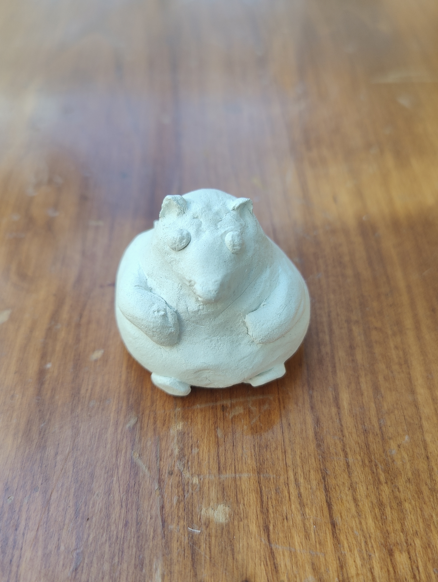 A small, roughly spherical, clay figure resembling a stylized bear is centered on a polished wooden surface. The figure is light greenish-gray in color, with rudimentary details indicating eyes, ears, a snout, and paws. The wooden surface is light brown with a noticeable grain pattern running vertically in the frame. The scene is brightly lit, casting a soft shadow beneath the clay figure. There is no indication that the location is Ipswich, United Kingdom or any identifying characteristics of the scene.