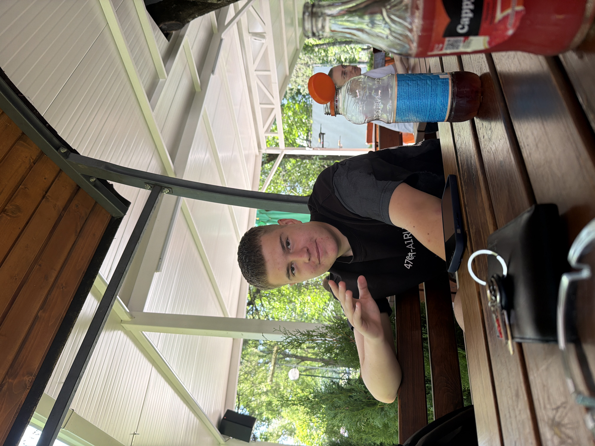 A young man with short, light brown hair and a light complexion is sitting at a wooden table outdoors. He is wearing a black t-shirt with a graphic print. His left arm is resting on the back of a wooden bench, and his right hand is raised with fingers splayed. He is looking towards the camera with a slight smile. On the table in front of him is a black wallet with keys on top, a dark-colored smartphone, and two glass bottles. One bottle is clear with a blue and white label and an orange cap, and the other is red with a label that is partially visible. In the background, trees and foliage are visible, suggesting an outdoor setting in a park or garden. The overall environment appears to be a covered seating area, with a wooden roof and white ceiling panels.