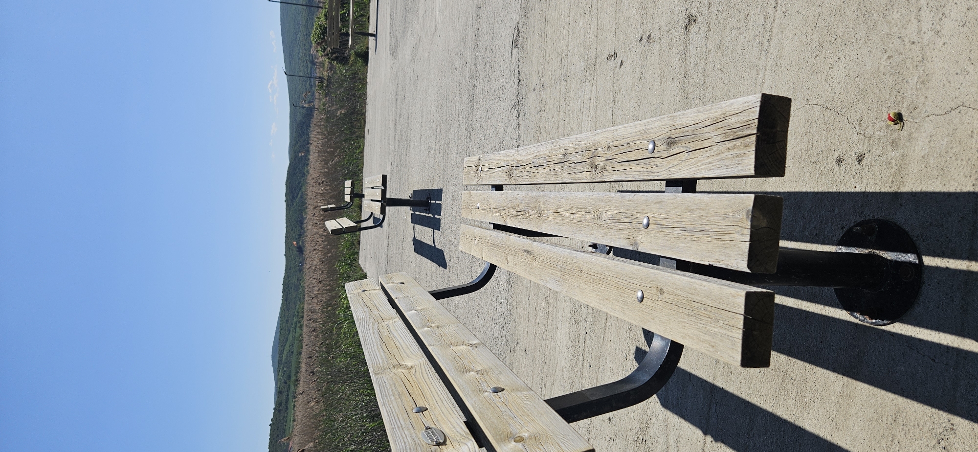 The image shows a row of wooden benches on a concrete platform. The benches are light-colored wood with metal supports. The landscape behind the benches features vegetation and a clear blue sky. In the upper right corner, there is a small red object on the concrete. The scene is located in Δημοτική Ενότητα Προμαχώνος, Greece.