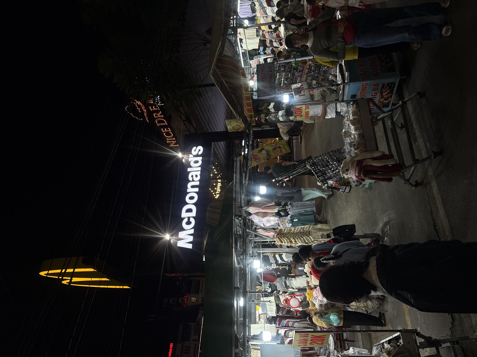 This is a nighttime street scene in Da Lat, Vietnam, with a prominent McDonald's sign illuminating the area. The foreground shows a bustling night market with various stalls selling clothing and food. Several people are browsing the merchandise and interacting with vendors. In the background, the McDonald's restaurant is visible, as is a sign for "NICE DREAM." The overall atmosphere appears lively and energetic, typical of a Vietnamese night market.