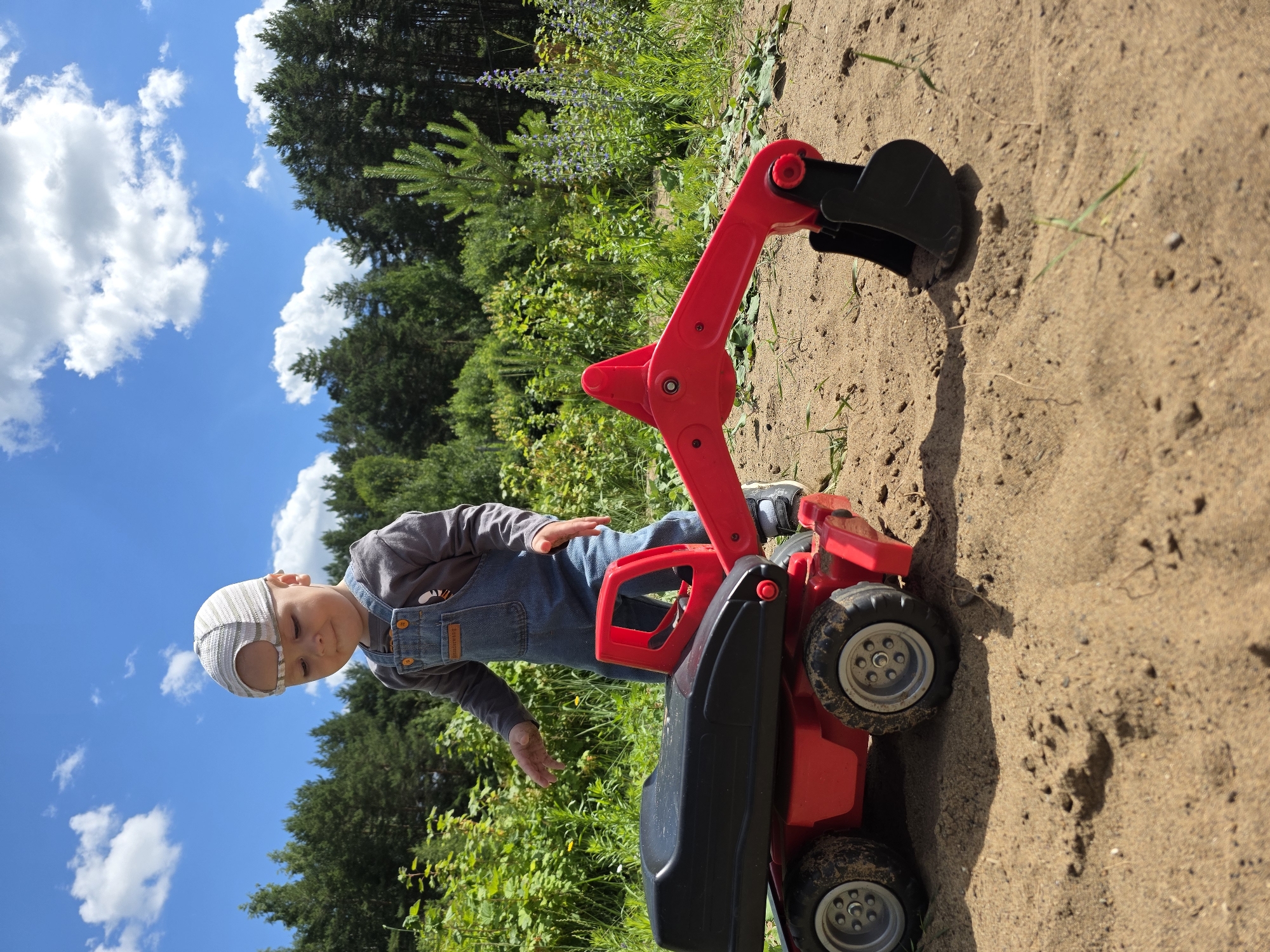 A young child is playing with a red toy excavator in a sandy area outdoors. The child is wearing denim overalls, a grey long-sleeved shirt, and a striped cap. They are leaning towards the toy, with their hands outstretched as if operating it. The toy excavator is large and red with a black bucket and wheels. The background consists of lush green trees and a bright blue sky with scattered white clouds. The scene appears to be set on a sunny day, likely during the daytime. The ground is sandy, with some patches of grass and small plants growing. The overall mood is one of playful activity and enjoyment in an outdoor environment. The location is identified as Chaikovsky, Russia.