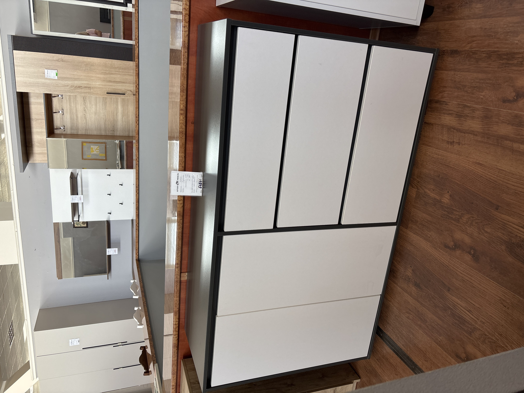 This media file displays a retail setting featuring furniture. The primary objects are a large, modern cabinet with white doors and a dark frame, and a hallway wardrobe with a light wood finish and a mirror. The hallway wardrobe is to the left of the cabinet and shows hooks for hanging clothes and a small framed picture above a mirrored section. The cabinet, positioned to the right, is larger and has multiple doors.
The scene is set indoors, likely in a furniture store or showroom, indicated by the display of furniture items and the presence of price tags. The floor is made of wooden planks. The lighting appears to be artificial, suggesting an indoor environment regardless of the time of day.
There are no people visible in the media file. The overall impression is one of a clean and organized retail space.
Visible text includes price tags on the furniture. On the hallway wardrobe, a tag reads "АРМАН МЕБЕЛЬ" and a price "6484". Another tag on the same wardrobe has smaller text that is difficult to read. On the cabinet, there is a visible tag with text that is also too small to decipher.