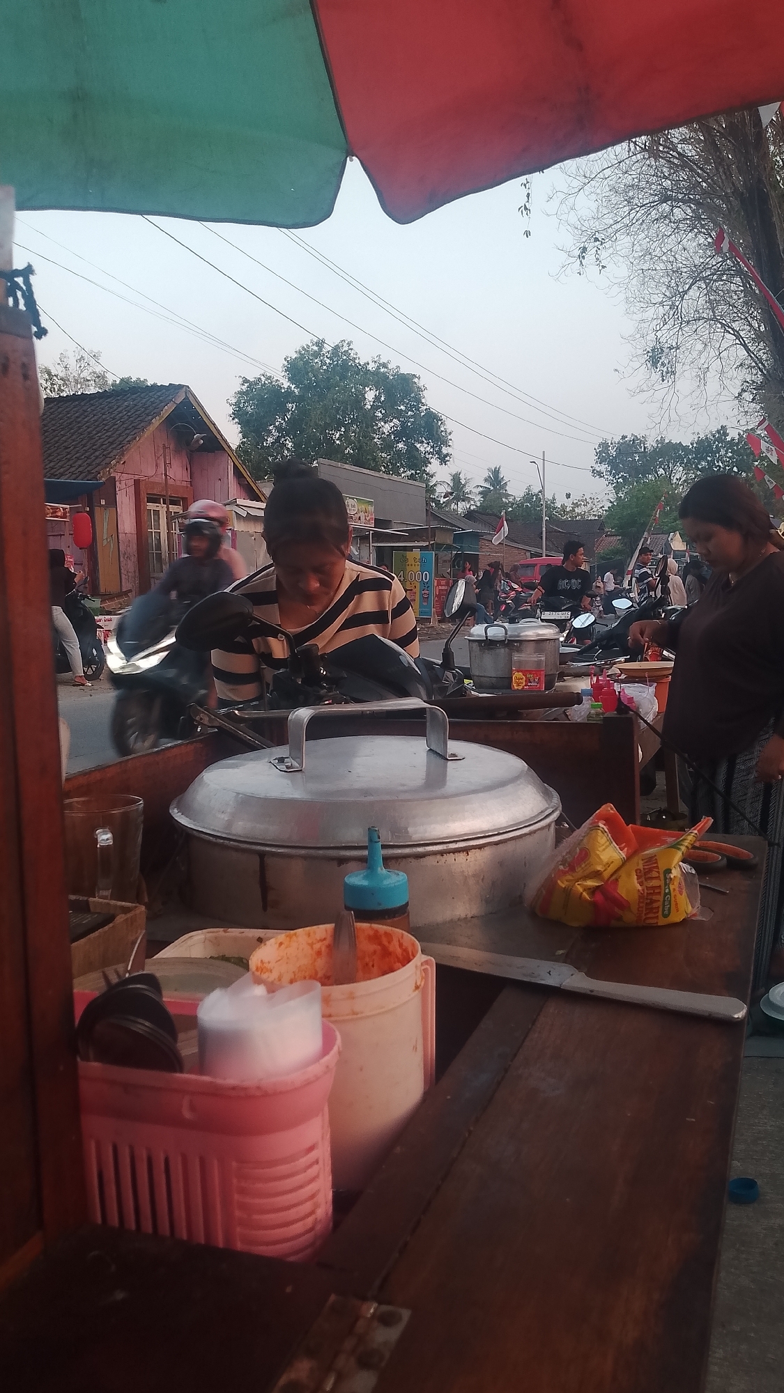 An outdoor street food stall in Karanglangu, Indonesia, features a woman in a striped shirt attending to a large metal pot with a lid. A second woman in a dark shirt stands to her right. In the foreground, various food preparation items are visible, including a pink utensil holder with white cups and a large white bucket containing red food and a utensil. A bag of snacks rests on the counter to the right. The background shows a street scene with motorcycles and buildings, including a pink building with a dark roof. The sky is overcast, and a green and red awning covers part of the stall.