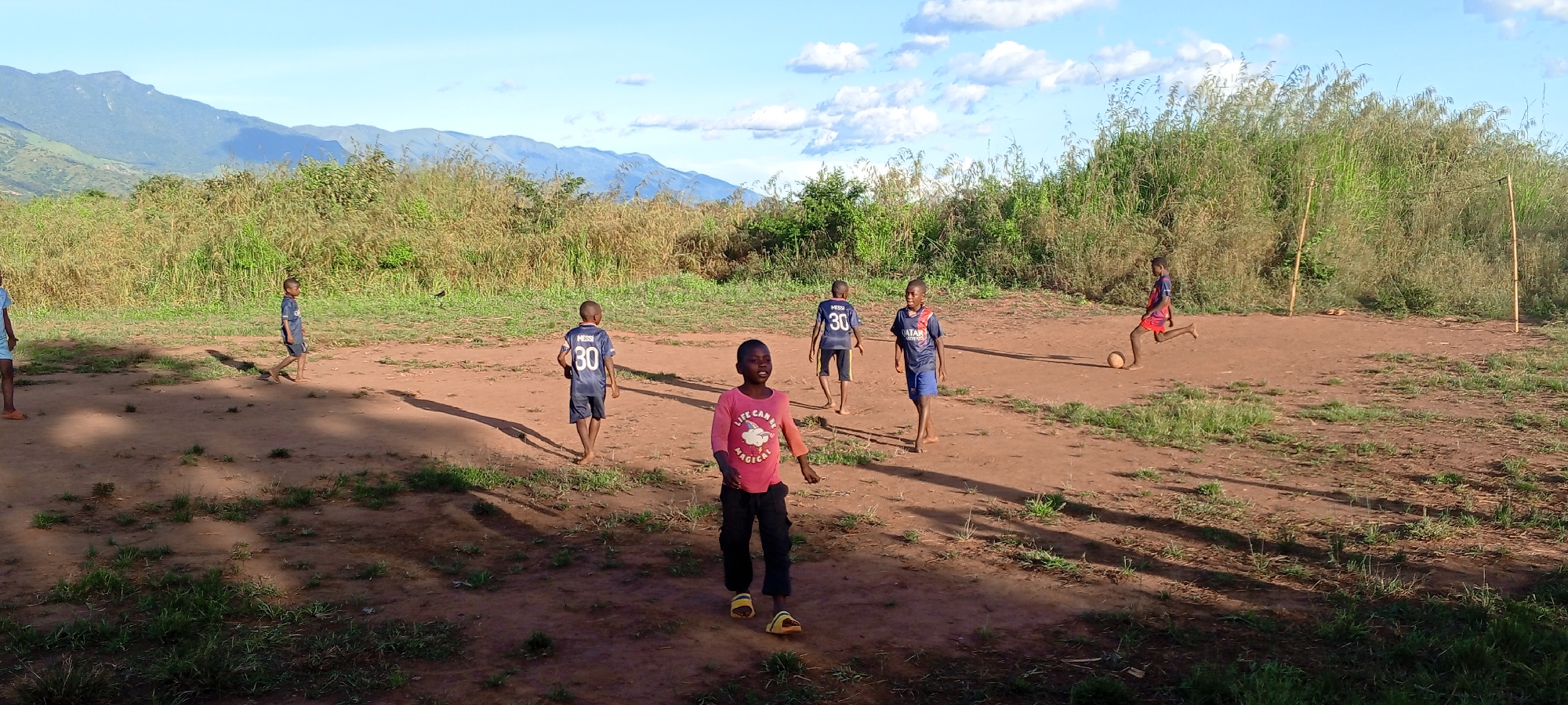 In Ngalula, Democratic Republic of the Congo, several children are playing soccer on a dirt field. In the foreground, a child in a pink shirt and dark pants walks toward the viewer. Behind him, other children wearing soccer jerseys are scattered across the field. One is kicking a soccer ball towards a goal made of wooden posts. Tall grass and vegetation surround the field. In the background are mountains under a partly cloudy sky.
