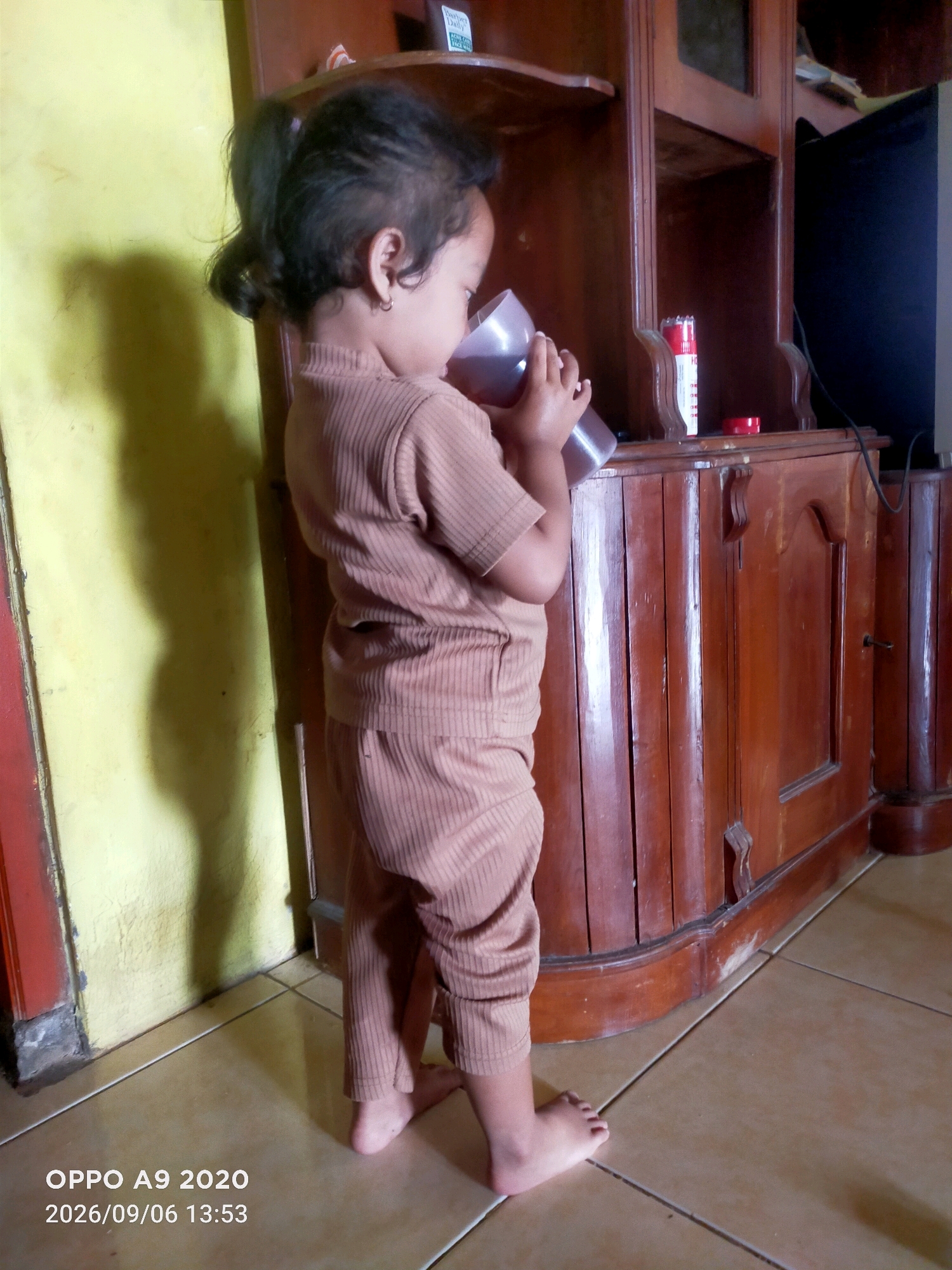 The image shows a young child standing on a tiled floor, wearing a tan ribbed outfit and holding a brown cup to their mouth. The child is positioned to the left of a large, dark wood cabinet with multiple shelves and doors. The cabinet appears to be made of polished wood, and some items are visible on the shelves, including a red and white can. To the left of the child is a yellow wall, casting a shadow of the child onto it. The floor tiles are a light brown and beige color, arranged in a grid pattern. The timestamp "OPPO A9 2020 2026/09/06 13:53" is visible in the bottom left corner, suggesting the photo was taken on September 6, 2026, at 1:53 PM. The overall setting suggests an indoor residential environment, possibly a living room or dining area.