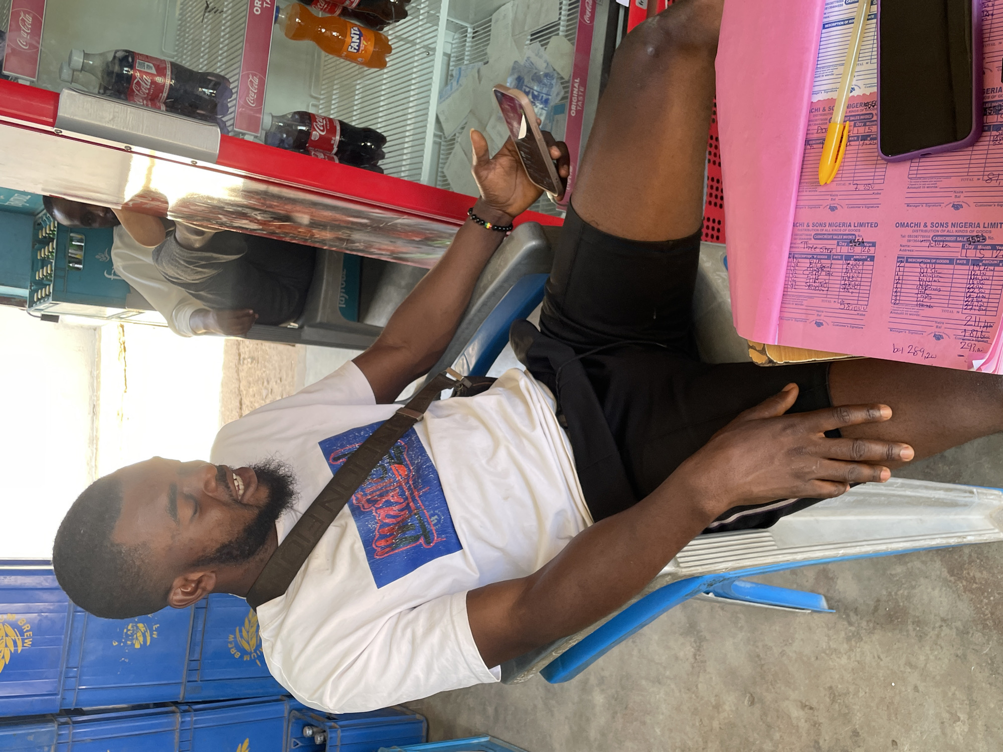 An eye-level shot captures a young Black man sitting on a blue and silver chair in what appears to be a shop or retail environment in Jos, Nigeria. He has a beard, short dark hair, and is wearing a white t-shirt with a blue and red graphic print, and black shorts. A dark strap from a bag runs across his chest. He holds a smartphone with a light-colored case in his right hand and has a beaded wristband on his left. The man has a slight smile, looking towards his phone or just past it.
Behind him and to his right, a large beverage refrigerator is visible, stocked with Coca-Cola, Fanta, and other bottled drinks. A bright reflection from overhead lighting or sunlight shines on the top of the refrigerator. To the man's left, stacked blue plastic crates, likely for beverages, are seen, with "BREW" text visible on some. On a surface to his right, a stack of pink documents, identified as "OMACHI & SONS NIGERIA LIMITED CASH/CREDIT SALES INVOICEs," are present, along with pens. Visible text on the invoices includes "DISTRIBUTION OF ALL KINDS OF GOODS," "Day 15 Month 26," "AMOUNT K," various handwritten figures like "58,000," "211,000," "293,000," and phone numbers such as "08036778660." The floor is concrete. The overall atmosphere suggests a casual daytime moment, possibly during a break or while waiting for a transaction.