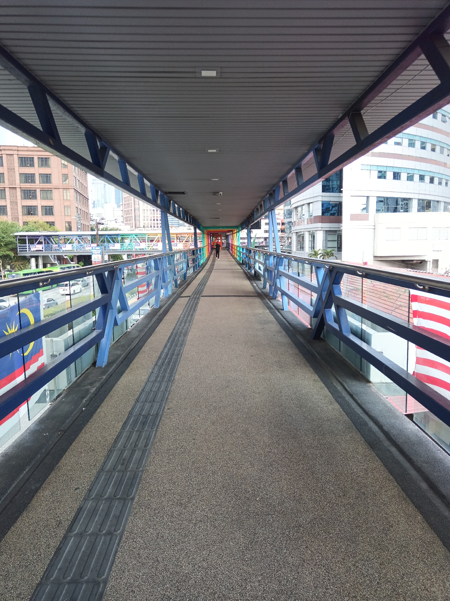 The image shows a perspective view of a pedestrian walkway or skybridge in Kuala Lumpur, Malaysia. The walkway is covered by a grey corrugated roof supported by dark blue metal beams. The walkway has a textured grey surface. Along the sides, there are blue railings with a criss-cross pattern, and clear glass panels below. On the left side of the walkway, there are flags resembling the Malaysian flag, with red and white stripes and a blue canton with yellow stars and crescent. On the right side, there are red and white striped banners. Beyond the walkway's railings, the urban environment of Kuala Lumpur is visible, including buildings with glass facades, trees, and a road with vehicles. In the distance, further along the walkway, other elevated pedestrian bridges are visible, some of which are decorated with colorful lights, possibly representing a rainbow. A single person, viewed from behind, is walking away from the camera on the walkway in the distance. The lighting suggests it is daytime, with diffused light.