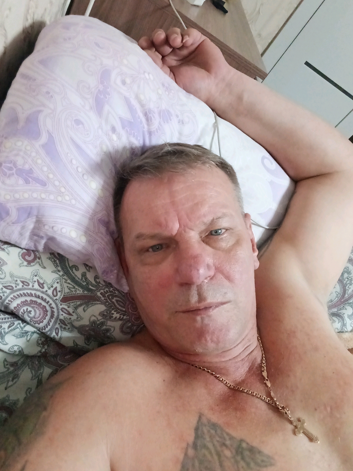 A close-up, overhead shot shows a middle-aged Caucasian male lying in bed. His head is propped on a pillow with a purple floral pattern. He has short, grey hair and is looking towards the camera with a neutral expression. He is shirtless, revealing a gold chain with a crucifix necklace and a tattoo on his chest. His right arm is bent and resting above his head, while his left arm is not fully visible. The background shows a bed with patterned sheets and a wooden surface that appears to be furniture. The scene is likely indoors, in a bedroom. There is no specific information about the location in Astrakhan, Russia.