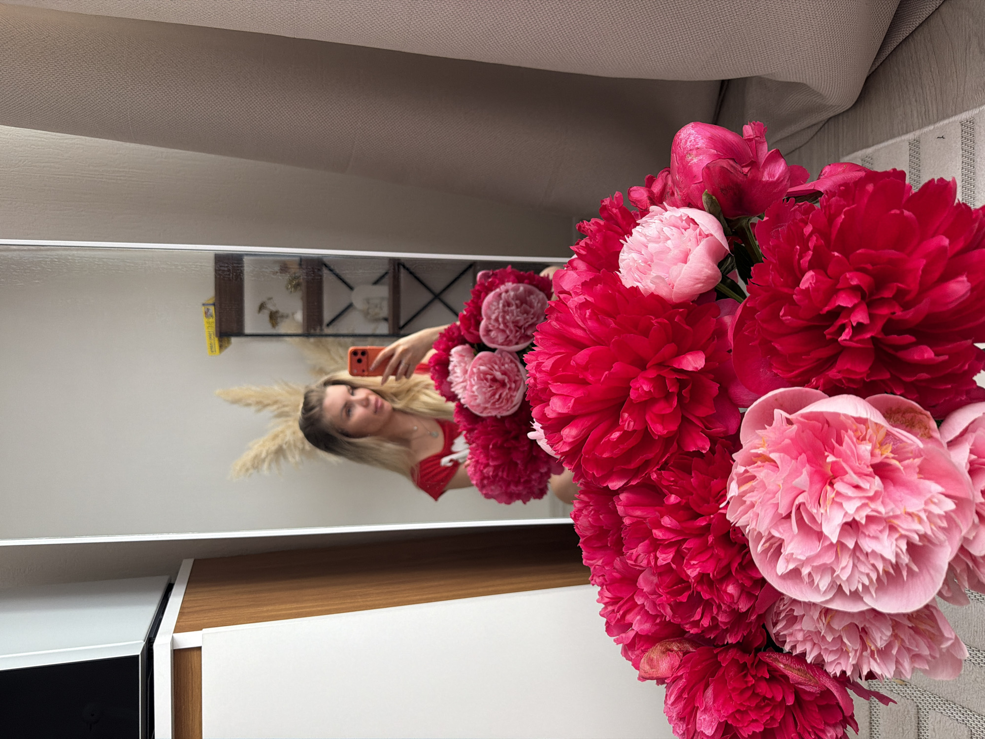 A woman with blonde hair is taking a selfie in a mirror, holding a red smartphone in her right hand. She is wearing a red t-shirt. In front of her, there is a large bouquet of pink and red peonies. The background includes a white and wooden cabinet, a mirror reflecting the scene, and some beige curtains. The setting appears to be an indoor residential space, possibly a bedroom or living room. There is no indication of the location being in Moscow, Russia.
