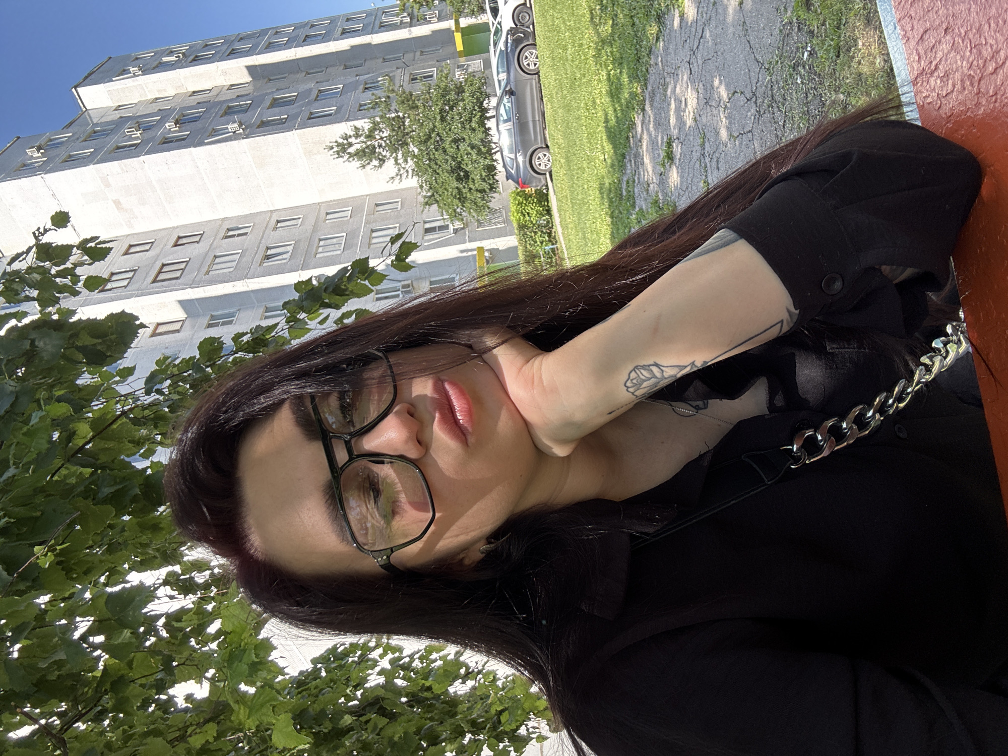 A young woman with long, dark hair and glasses is pictured from the chest up, looking at the camera with her hand propping up her chin. She is wearing a black shirt and appears to be sitting at a red table outdoors. Behind her, a tall, gray apartment building is visible, with trees and a bright blue sky in the background. Several cars are parked in front of the building.