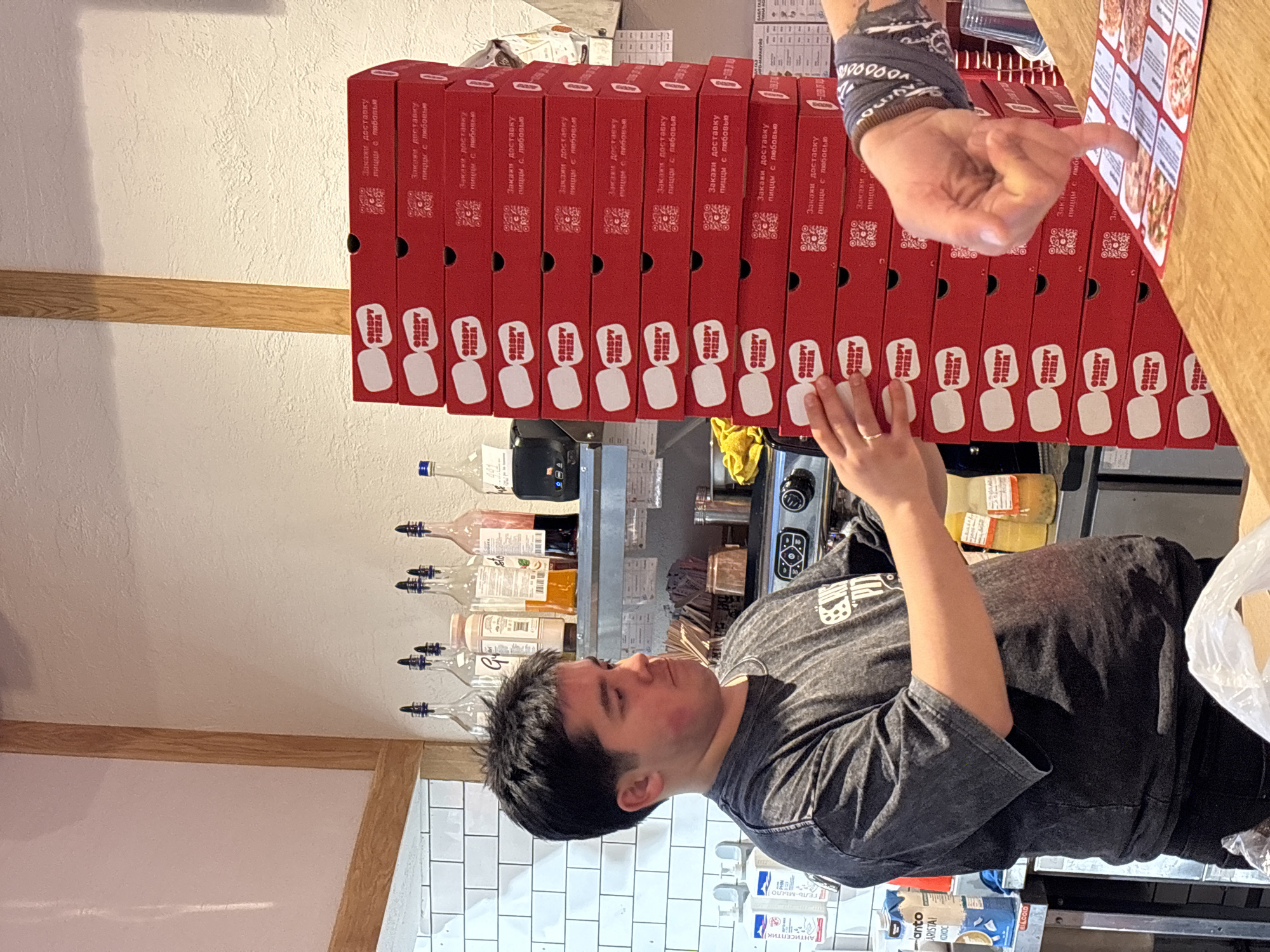 In Saint Petersburg, Russia, a young man with dark hair, wearing a grey t-shirt with a "CRISPY PIZZA" logo, stands behind a counter. To his right, a tall stack of red "CRISPY PIZZA" boxes is visible. A hand with a blue bandana and tattoos points at a menu on the counter in the foreground, where several pizza images are displayed. In the background, shelves hold bottles of syrups and other items. The environment appears to be a pizza shop.