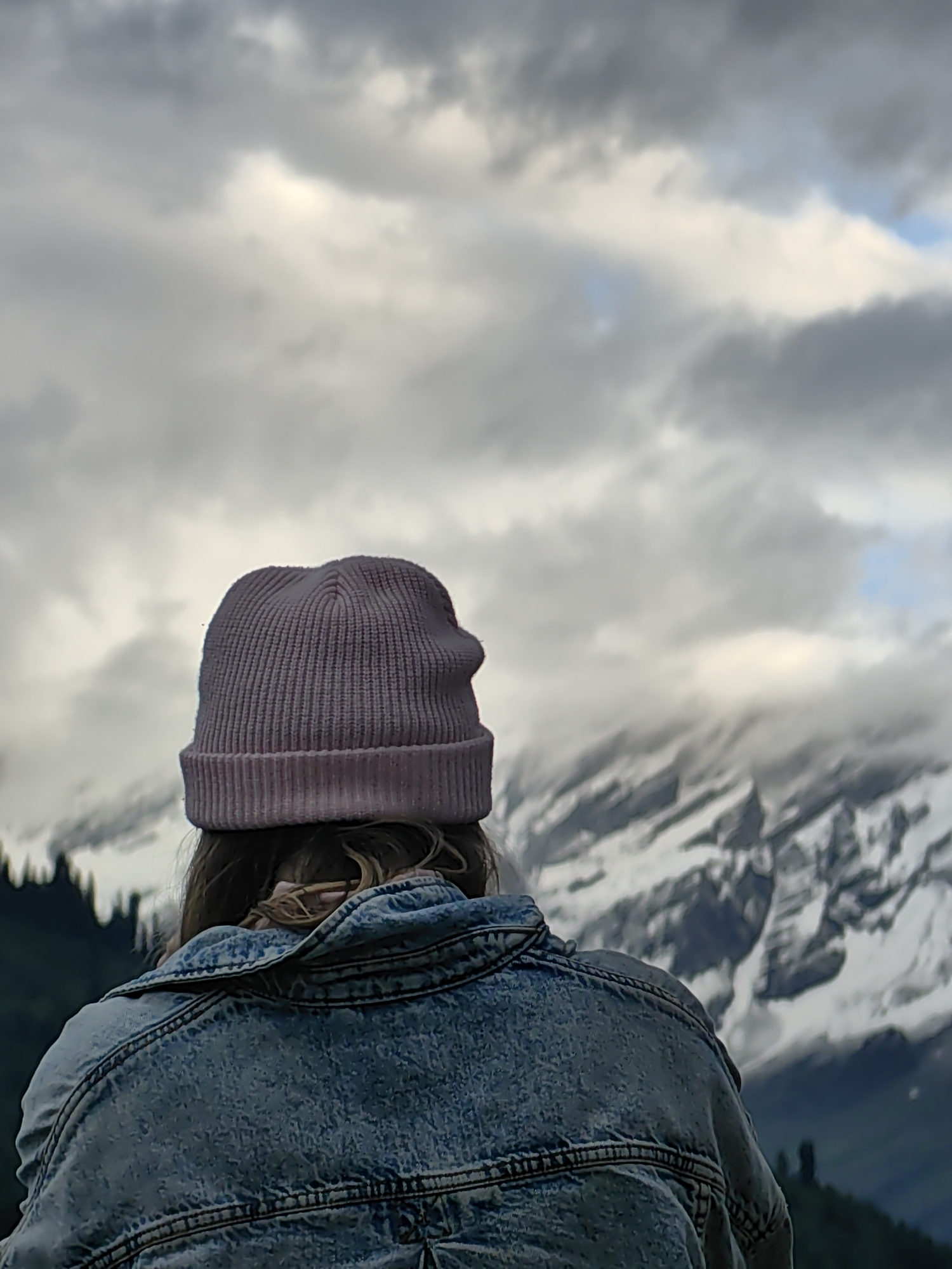 The image shows a person from behind, wearing a light pink knitted beanie and a blue denim jacket. They are looking towards a mountainous landscape under a cloudy sky. The mountains are covered in snow and appear rugged. The scene is likely an outdoor setting, possibly a viewpoint overlooking a valley or mountain range. The location is specified as Vashisht, India, a town known for its hot springs and proximity to the Himalayas. The lighting suggests it is daytime, but the clouds create a somewhat muted atmosphere. The focus is on the person and their gaze towards the expansive natural scenery.