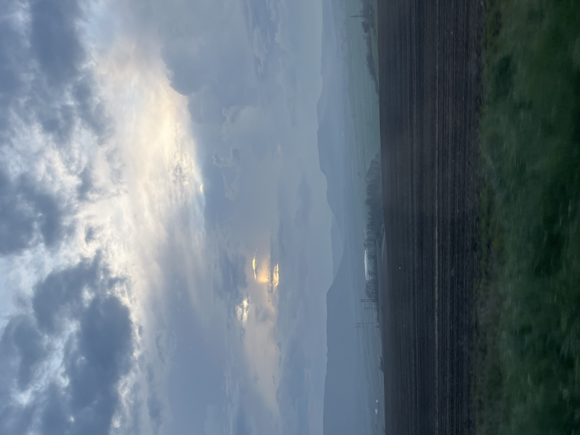 The image depicts a landscape under a cloudy sky, possibly Petrov Dol, Bulgaria. The sky is filled with various cloud formations, with patches of sunlight breaking through. Below the clouds, there is a distant mountain range, partially obscured by haze or rain. The foreground consists of a cultivated field with dark soil and a strip of green grass or vegetation on the right. Power lines are visible. The image has a slightly blurred quality, possibly due to motion or atmospheric conditions.